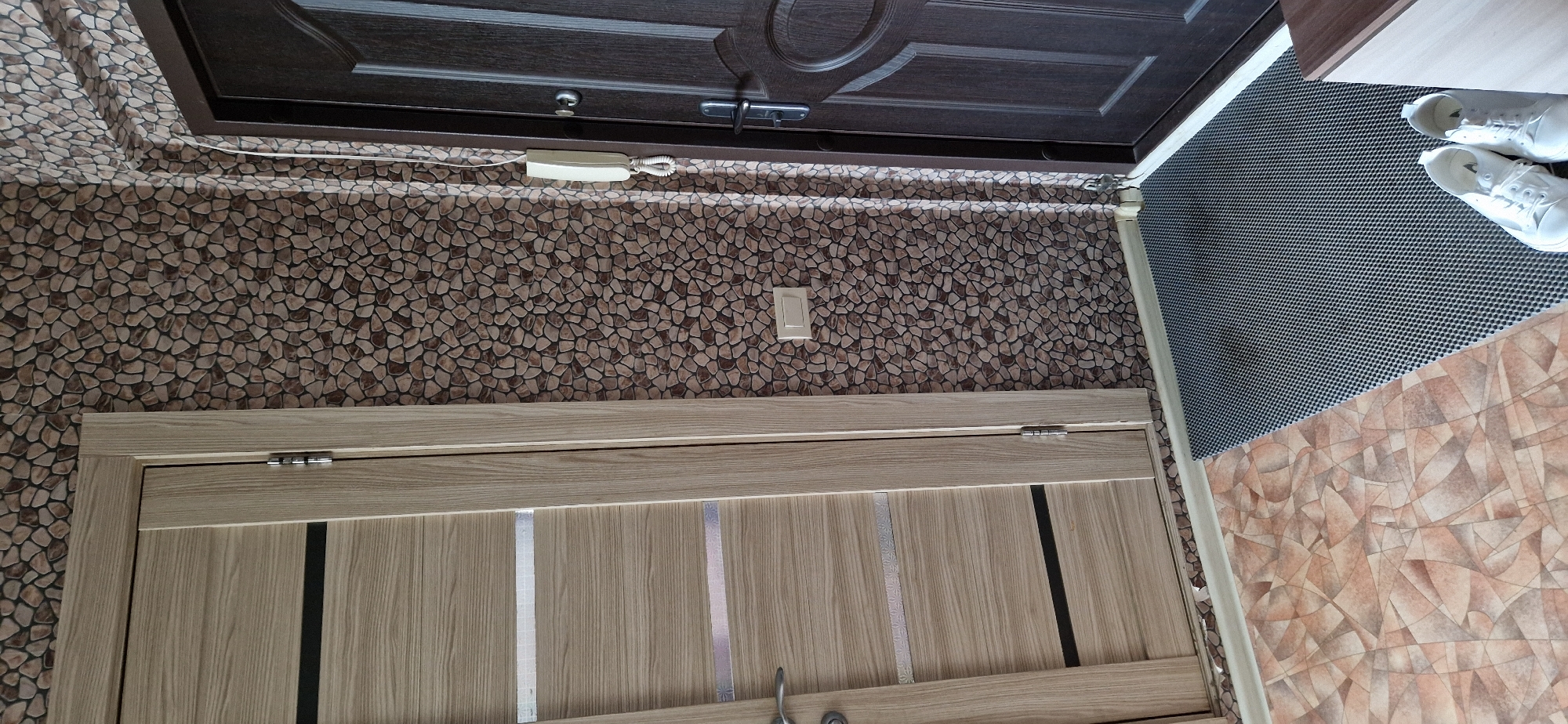 A close-up, eye-level shot shows a hallway with two doors and a textured wall. The left door is a light wood grain with two horizontal black inserts and a silver decorative strip above and below the lower black insert. The right door is a dark brown, possibly metal, with a rounded design element. Between the doors, the wall is covered in wallpaper with a pattern of small, irregular brown and beige stones. A white intercom unit is mounted on the wall to the right of the light-colored door and above a small rectangular light switch. The floor is covered in two types of flooring: to the left, a patterned tile with geometric shapes in shades of orange, beige, and pink, and to the right, a dark gray rubber mat with a hexagonal pattern. A pair of white canvas sneakers is placed on the gray mat in the lower right corner of the image. The scene appears to be an interior hallway, possibly near an entrance.