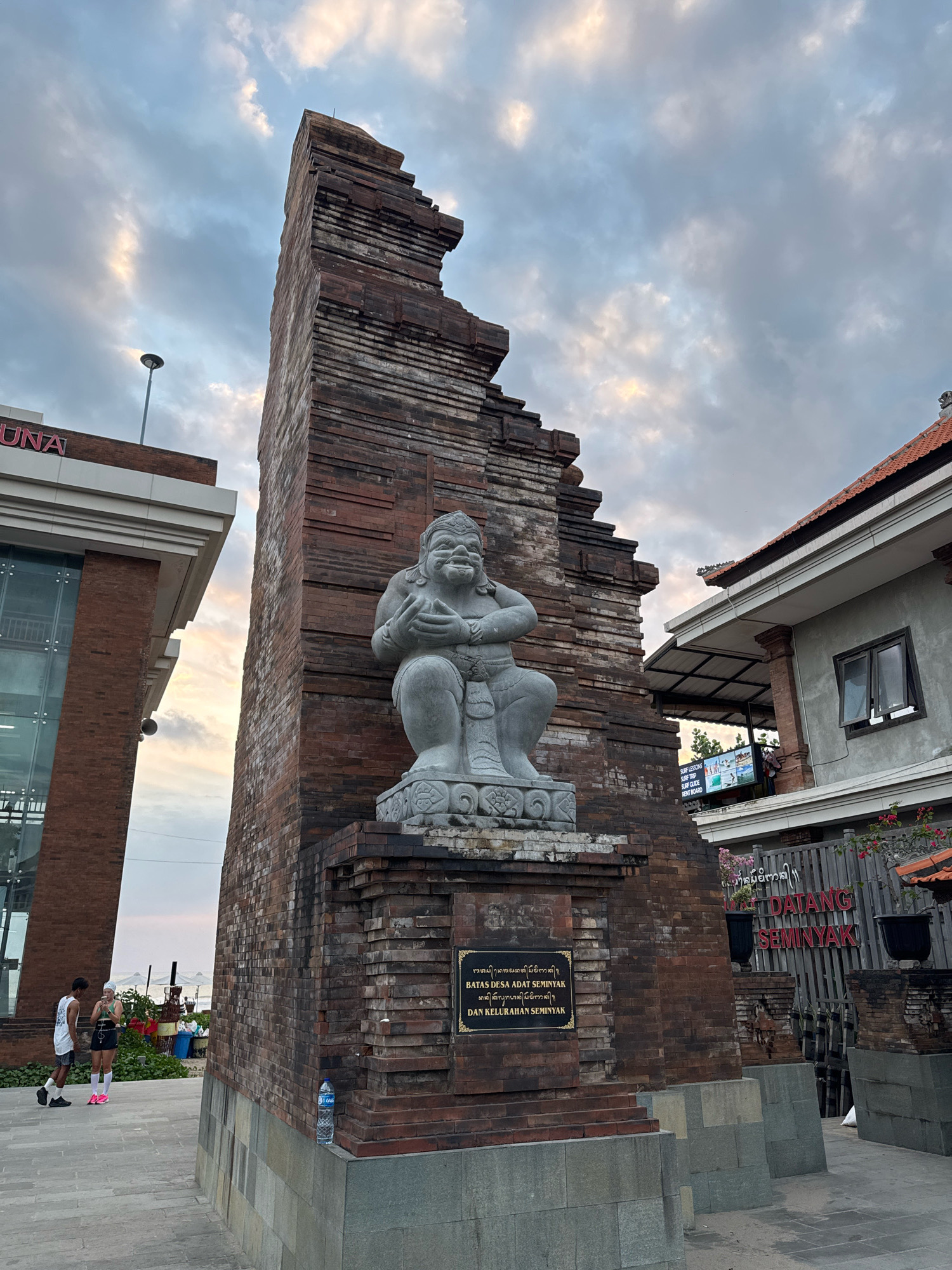 The image depicts an outdoor scene in Seminyak, Kuta, Indonesia, likely at dusk given the lighting and cloudy sky. The central focus is a tall, weathered brick structure that appears to be an ornamental gateway or monument. A stone statue of a seated, robust figure with a benevolent expression is positioned on a pedestal in front of the brick structure. A dark plaque with text in an unfamiliar script is attached to the base of the structure.
To the left of the brick monument is a modern building with a glass facade and brick accents. The word "UNA" is visible in red lettering on the side of this building. Two individuals, a man and a woman, are standing on a paved walkway in front of this building, appearing to be engaged in a conversation. They are dressed in casual athletic wear.
To the right of the brick monument, a building with a traditional tiled roof and grey facade is visible. A sign with the words "DATANG SEMINYAK" in large red letters is mounted on a wooden fence. Below this sign, a smaller sign displays information about surf lessons, surf guides, and rentals. Potted plants adorn the area.
Further in the background, beyond the immediate structures, hints of a beach or coastal area can be discerned with what appear to be beach umbrellas and people. A plastic water bottle is placed on the base of the brick structure. The overall atmosphere suggests a public space near a popular tourist destination.