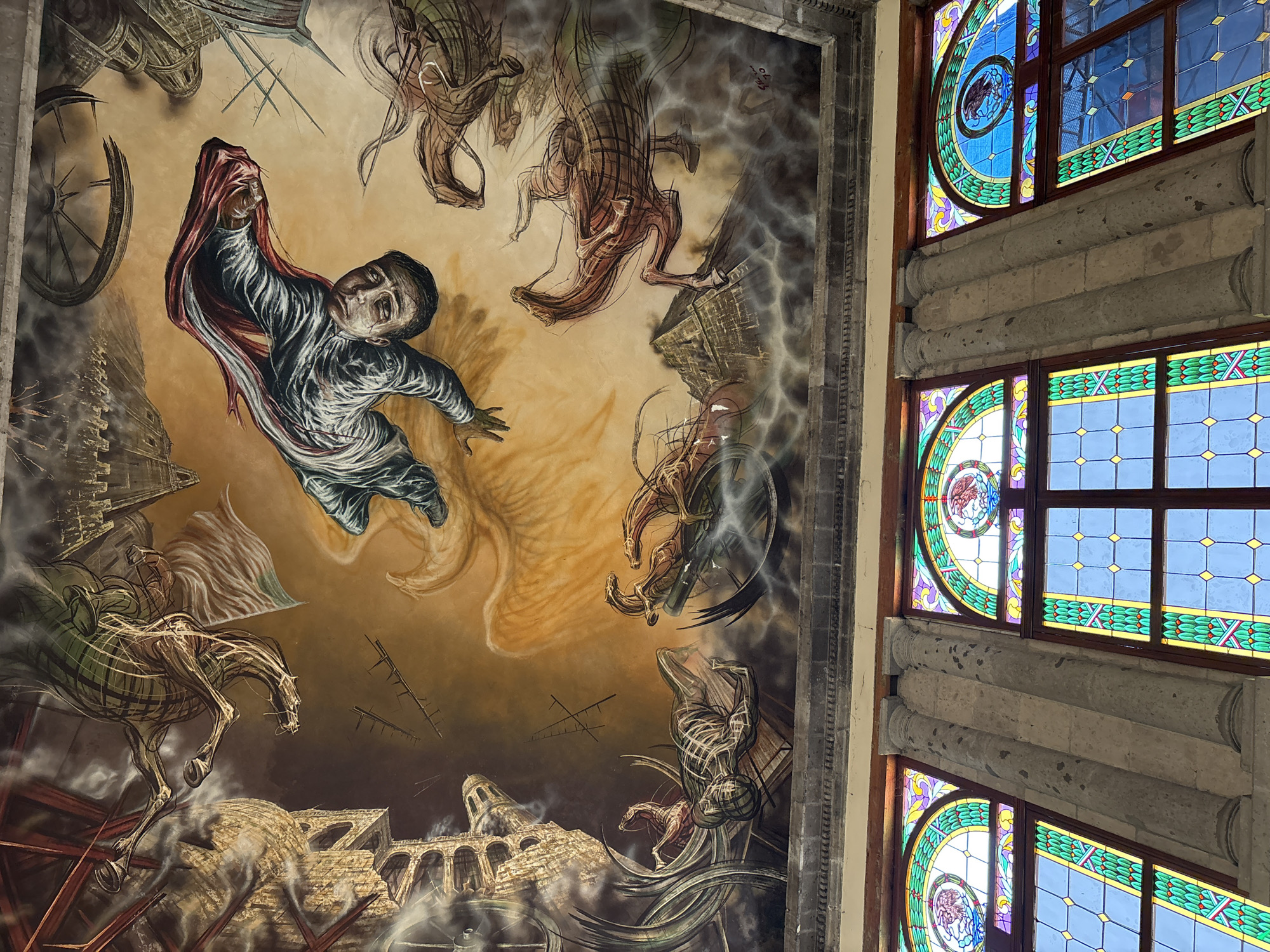 The image shows a portion of a mural painting on a ceiling, alongside stained glass windows. The mural depicts a chaotic scene of destruction with distressed figures, skeletal horses, and crumbling architecture, possibly representing a historical event or allegorical representation of war or disaster. One central figure appears to be falling or being thrown, partially covered by a red cloth.
The stained glass windows are multicolored, with geometric patterns and circular elements containing what appears to be an eagle emblem, possibly indicating the location as a governmental or institutional building in Mexico.
Based on the imagery and location, it is likely that this image captures a segment of Diego Rivera's mural "México de Hoy y de Mañana" located at the National Palace in Mexico City, Mexico.