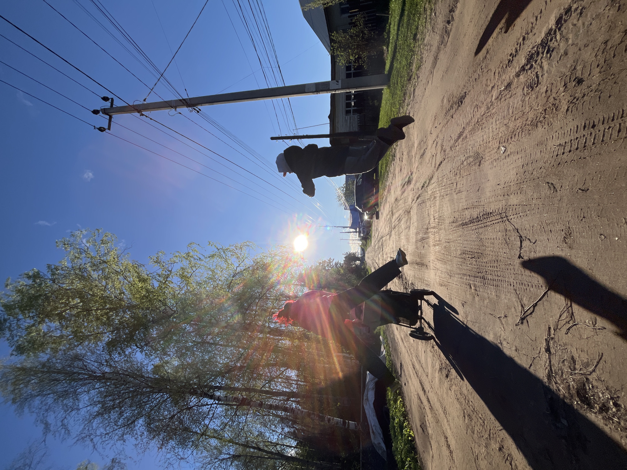 The image shows a dirt road in Chaikovsky, Russia, with tire tracks visible. A person is walking on the road with a stroller, accompanied by a dog. A second person is walking further away down the road. Power lines and a utility pole are visible against a blue sky. There are trees to the left and a building in the background. The sun is shining brightly, creating lens flare in the image.