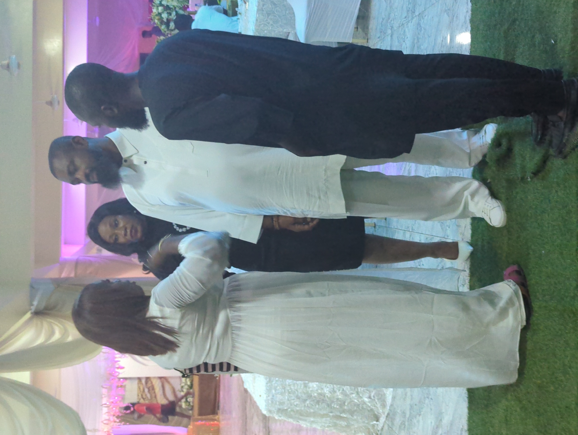 The image captures a social gathering at an indoor venue in Barden Yana, Nigeria. Four individuals are visible. On the left, a woman in a long, white dress is seen from the waist down, her feet clad in pink sandals. To her right, a woman in a black dress stands with her arms partially visible. Center-left, a man with a beard, wearing a white tunic and trousers, faces forward, engaged in conversation with the man to his right. The man on the right, with a beard, wears a black outfit and is seen from the shoulders down, facing left. The environment features draped fabric decorations in the background and tables covered with white cloths. The floor is partially covered with green artificial turf.