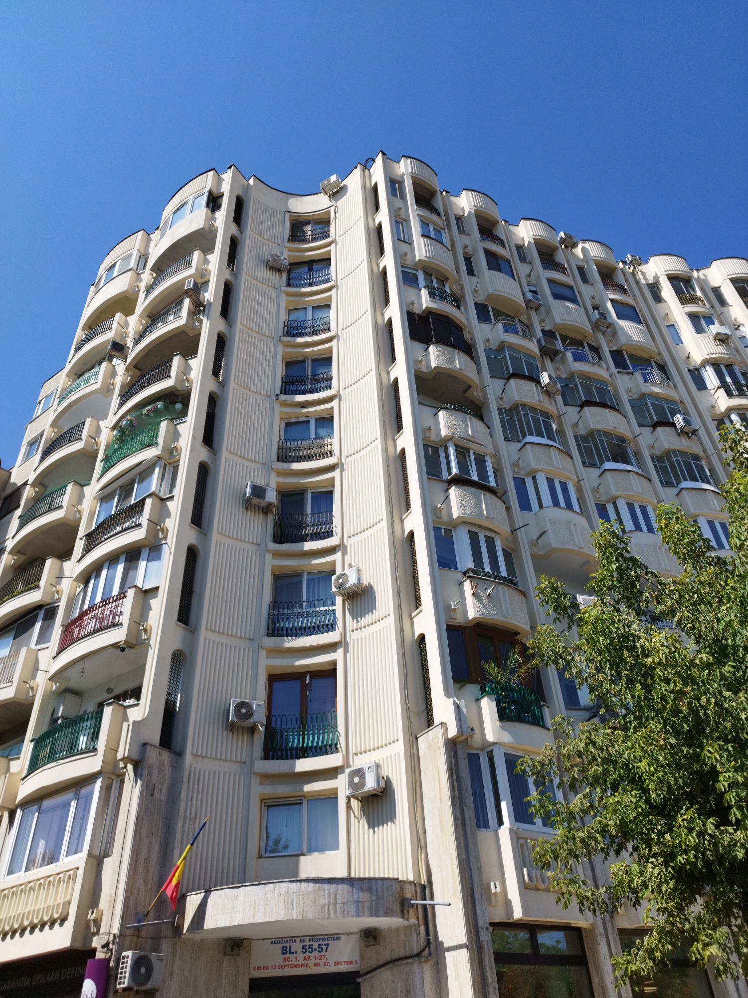 The image shows a tall, cream-colored apartment building with a curved facade, seen from a low angle against a clear blue sky. Numerous balconies, many with dark metal railings and some with decorative green fencing, protrude from the building. Various air conditioning units are visible mounted on the exterior walls. A Romanian flag, partially unfurled, is attached to the building near the ground floor entrance. Below the flag, a sign indicates "ASOCIATIA DE PROPRIETARI BL. 55-57 SC. 1, AP. 1-27 CALEA 13 SEPTEMBRIE, NR. 57, SECTOR 5". This indicates the building is located at Calea 13 Septembrie, No. 57, Block 55-57, Staircase 1, Apartments 1-27, in Sector 5 of Bucharest, Romania. Lush green foliage from a large tree partially obscures the lower right side of the building. The overall impression is of a residential building in an urban environment on a sunny day.