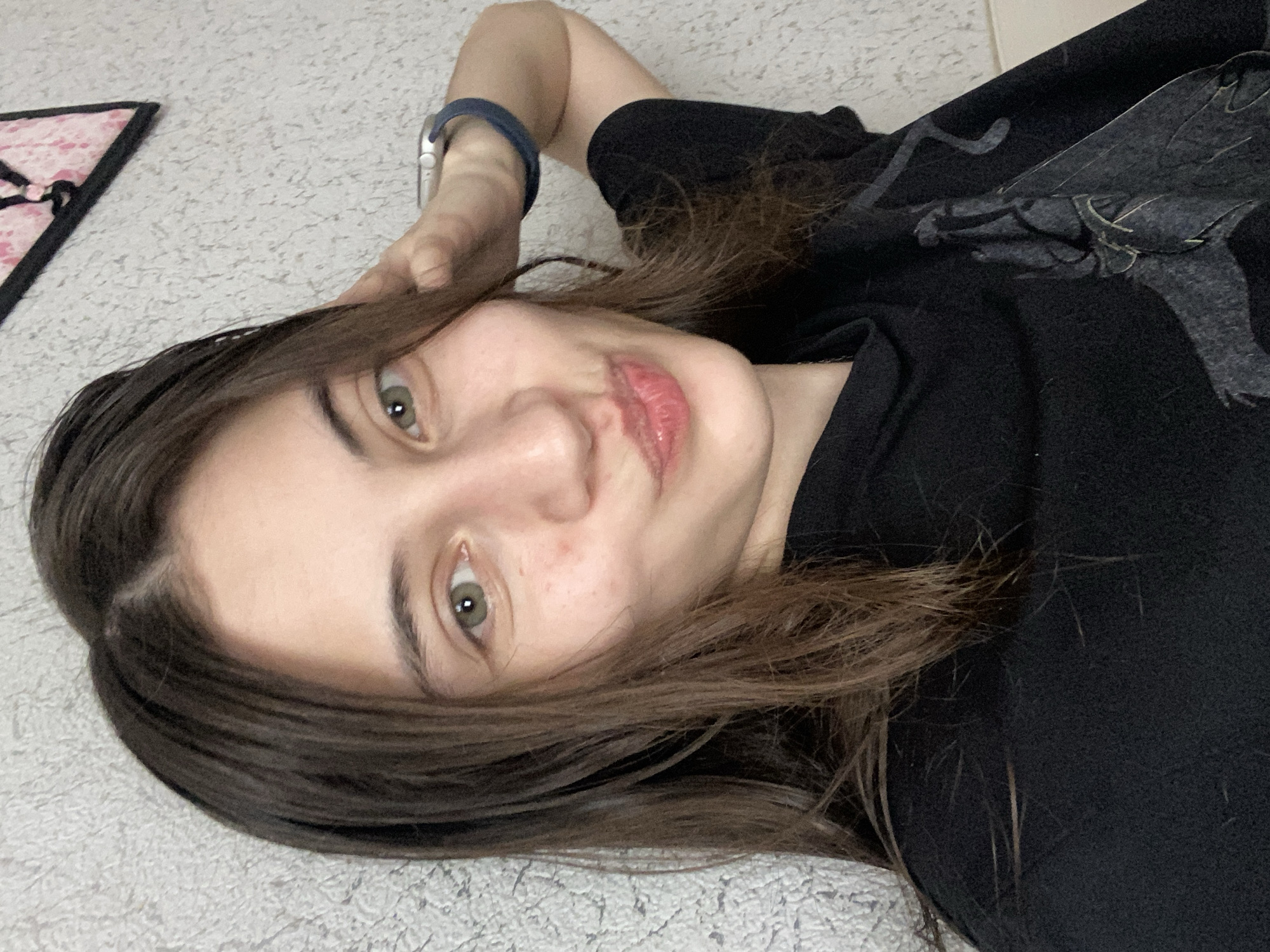 A young woman with long, dark brown hair and light green eyes is taking a selfie. She is wearing a black t-shirt with a graphic design. Her left hand is resting on the back of her head, and she is wearing a watch on her left wrist. Her lips are pursed, and she appears to be looking at the camera. The background is a textured, light-colored wall, with a pink and black triangular object hanging on it. The scene suggests an indoor setting, possibly a home, in Votkinsk, Russia.