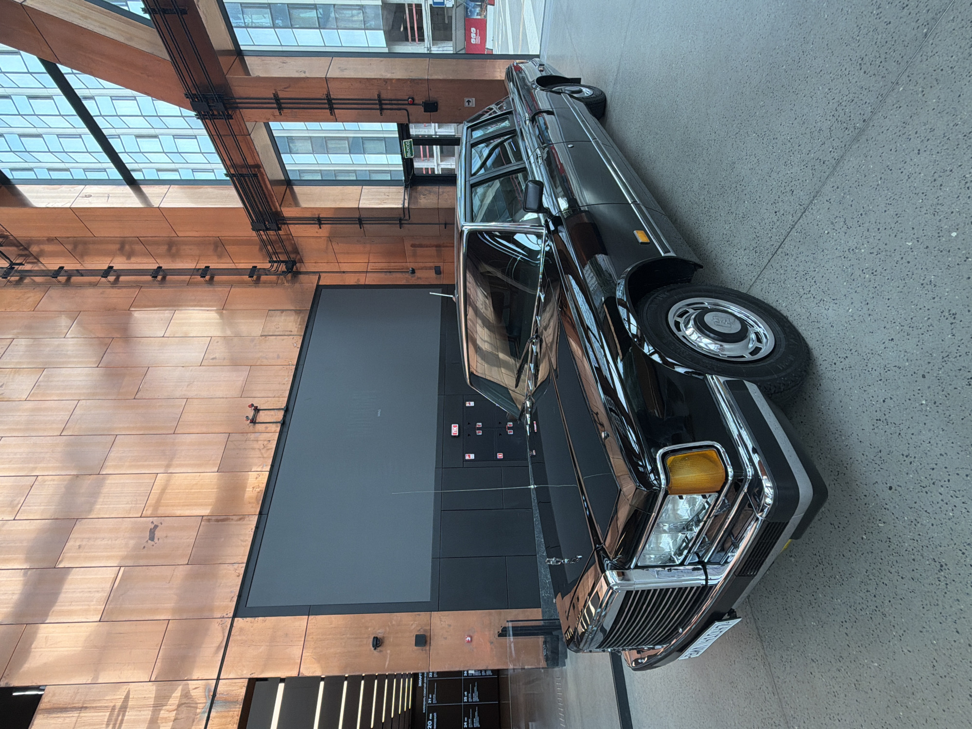 The photograph captures a black ZIL-4104 limousine parked indoors, likely in a public exhibition space or a modern building lobby in Moscow, Russia. The vehicle is positioned at an angle, showcasing its long, polished body and chrome detailing.
The environment features a backdrop of contemporary architecture. The walls are clad in large, rectangular copper panels with a brushed finish, exhibiting varying tones of brown and orange due to light reflection. To the right, a large window or glass facade reveals the exterior of a modern building with a grid-like structure of windows. Structural elements, possibly made of wood and metal, are visible, supporting the building's facade. Electrical conduits and wiring are neatly organized along the copper walls, indicating a functional interior.
In the background, a large, dark screen or digital display is mounted on the wall, its surface appearing blank or off. To the left, a section of an interior corridor with horizontal lighting fixtures is visible. Several signs are present, including a green "EXIT" sign with Cyrillic lettering ("ВЫХОД") above a doorway. Fire alarm boxes and other utilitarian elements are also integrated into the wall design.
The floor is a polished concrete surface with a speckled gray appearance, reflecting the overhead lighting and the car. The overall impression is one of a clean, modern, and well-lit interior, contrasting with the classic elegance of the Soviet-era limousine. No people are visible in the frame. The lighting appears to be a combination of natural light from the windows and artificial lighting from the ceiling and wall fixtures, creating a warm and somewhat dramatic ambiance.