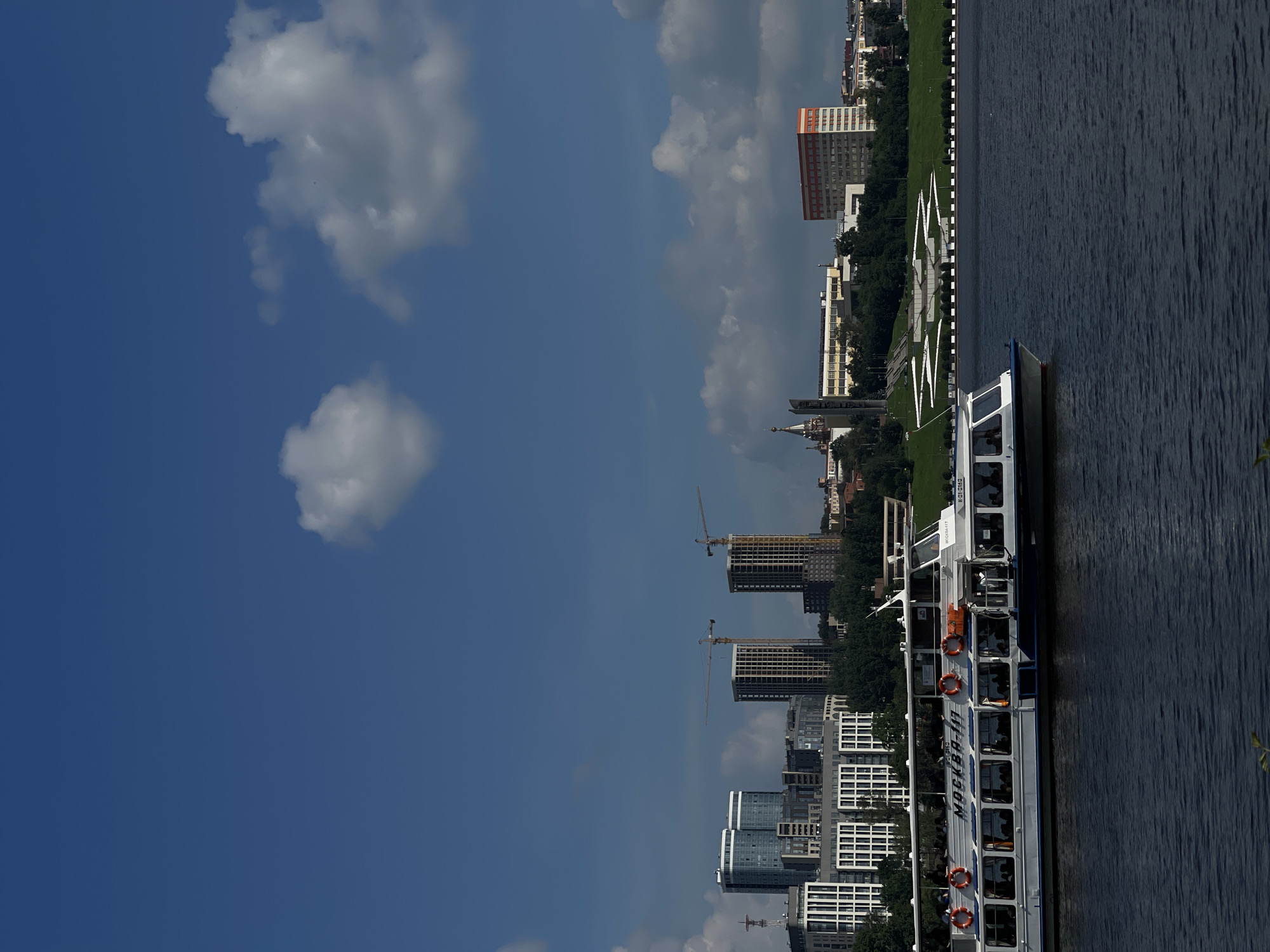 The image captures a wide-angle view of the Izhevsk city skyline from the waterfront. In the foreground, a white passenger boat with a blue stripe sails on a dark blue body of water. The boat has multiple windows revealing passengers inside. The word "МОСКВА-117" is visible on the side of the boat. In the middle ground, the Izhevsk skyline features a collection of modern buildings, including tall skyscrapers under construction with visible cranes. To the right, an orange and white rectangular building stands out. Behind the buildings, lush green trees line the embankment. The sky is a vibrant blue with scattered white clouds. A prominent feature of the embankment is a large, modern white structure with a repeating geometric pattern, resembling a stylized flower or star.