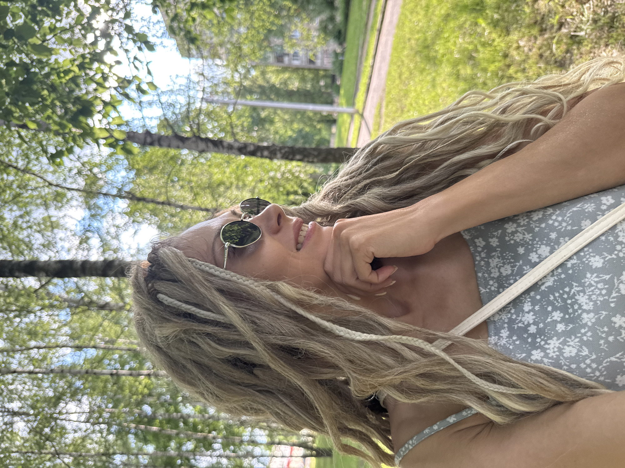 In an outdoor setting, a woman with blonde dreadlocks and sunglasses is captured from the chest up, looking to her right. She is wearing a light blue dress with a floral pattern and a white strap across her chest. Her right hand is resting on her chin, and she appears to be smiling. The background is blurred but shows trees with green foliage, suggesting a park or a tree-lined street in a city. Sunlight filters through the leaves, creating a dappled effect. The overall impression is one of a casual, sunny day outdoors.