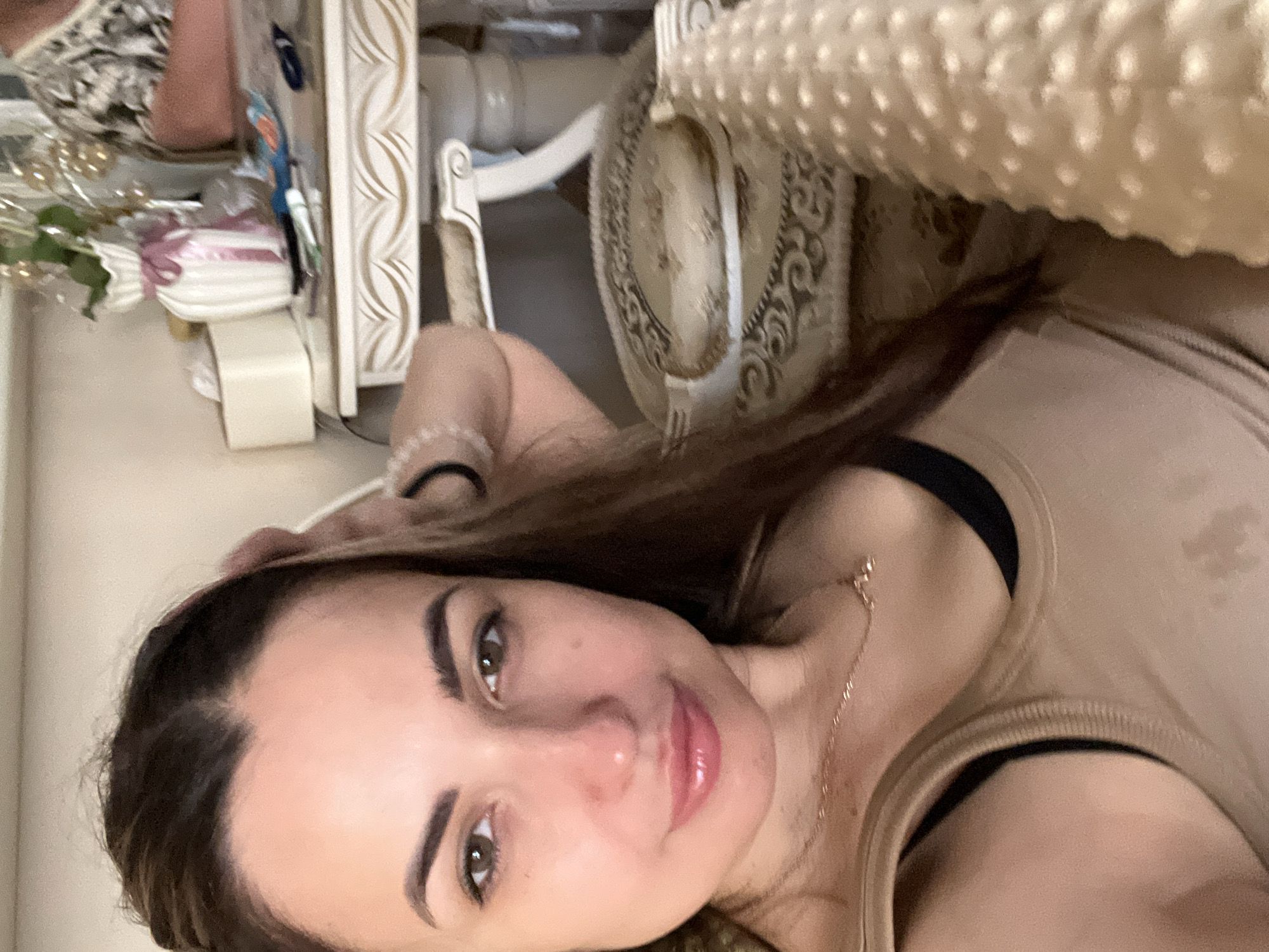An overhead selfie captures a woman in her late 20s to early 30s from the chest up. She has long, dark brown hair and is wearing a tan tank top with a black bra underneath. Her left hand is raised and touching her hair, while her right arm is bent, resting behind her head. She is looking directly at the camera with brown eyes, a neutral expression, and glossy pink lips. A thin gold necklace is visible around her neck.
The background is softly blurred and appears to be an indoor setting, possibly a living room or bedroom. To the right of the woman, a table with ornate carvings and decorative elements is visible. On the table, there's a white vase with a pink ribbon and what appears to be floral arrangements, along with a tissue box and some small objects. A portion of a chair with patterned upholstery is also in the frame. The lighting is soft and diffused, casting a warm glow on the scene.