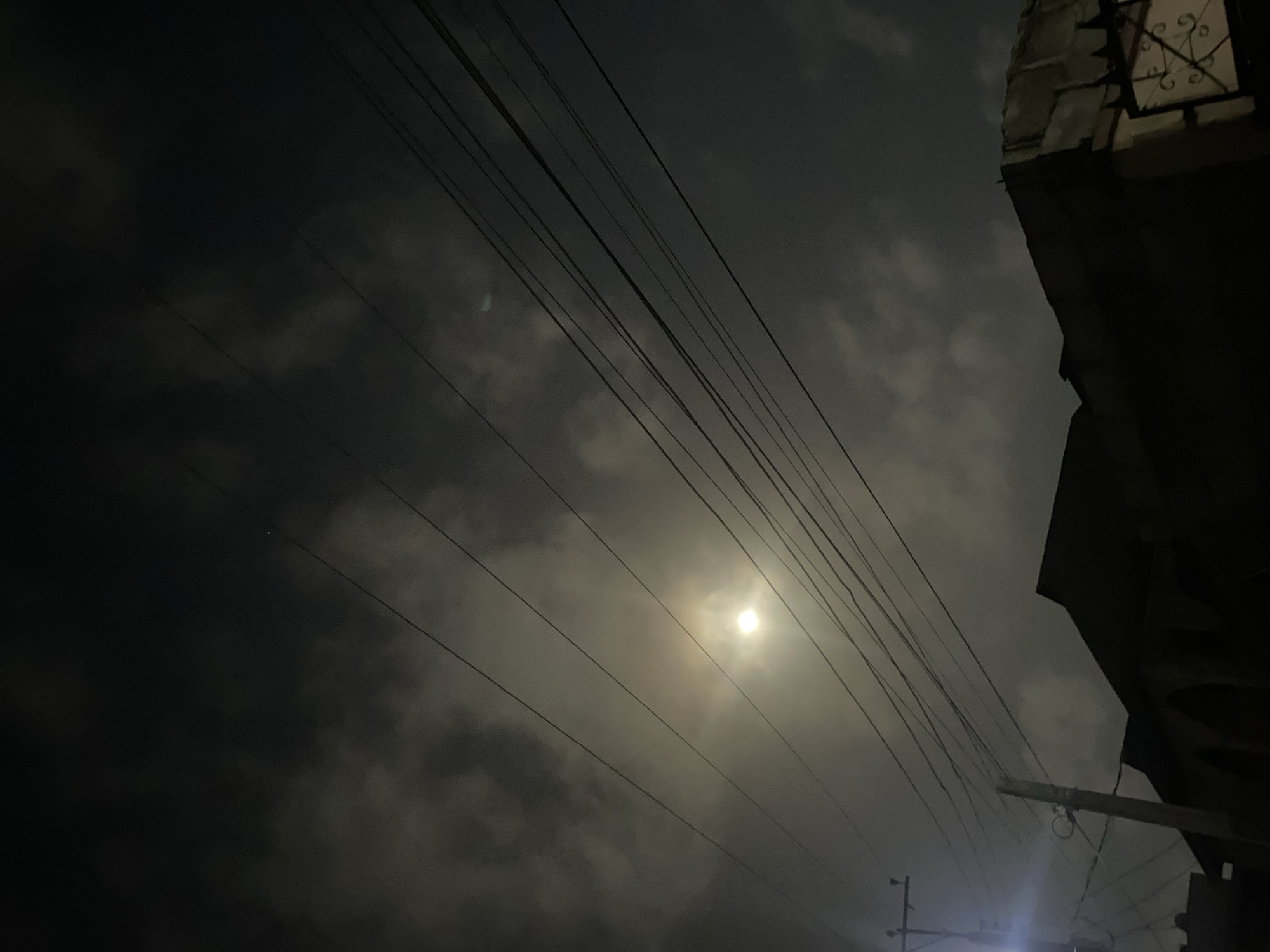 The image shows a night scene in Lagos, Nigeria. Overhead, power lines stretch diagonally across the frame, intersecting a cloudy sky where a bright, diffused light source is visible, likely the moon. To the right, a dark building occupies the upper corner, with a partial window grill visible. In the lower right, a utility pole stands, emitting a faint glow from a nearby light fixture. The general environment appears dimly lit. No people are visible.