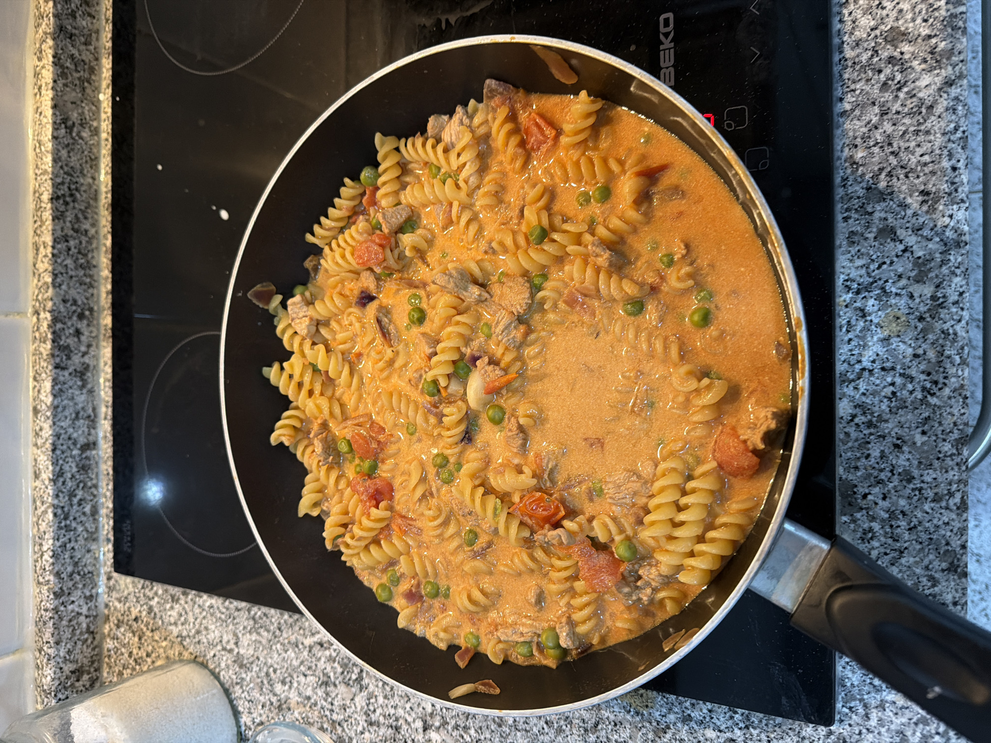 A top-down, eye-level shot depicts a skillet filled with cooked pasta, peas, meat, and tomatoes in a pink sauce, situated on a black glass stovetop. The stovetop is part of a counter with a grey speckled pattern. The handle of the skillet is visible on the right, made of silver and black material. The stovetop also features white markings and touch controls. The image suggests the scene is indoors, likely a kitchen, but the specific location in Vila Real, Portugal, cannot be determined from the visual information provided.