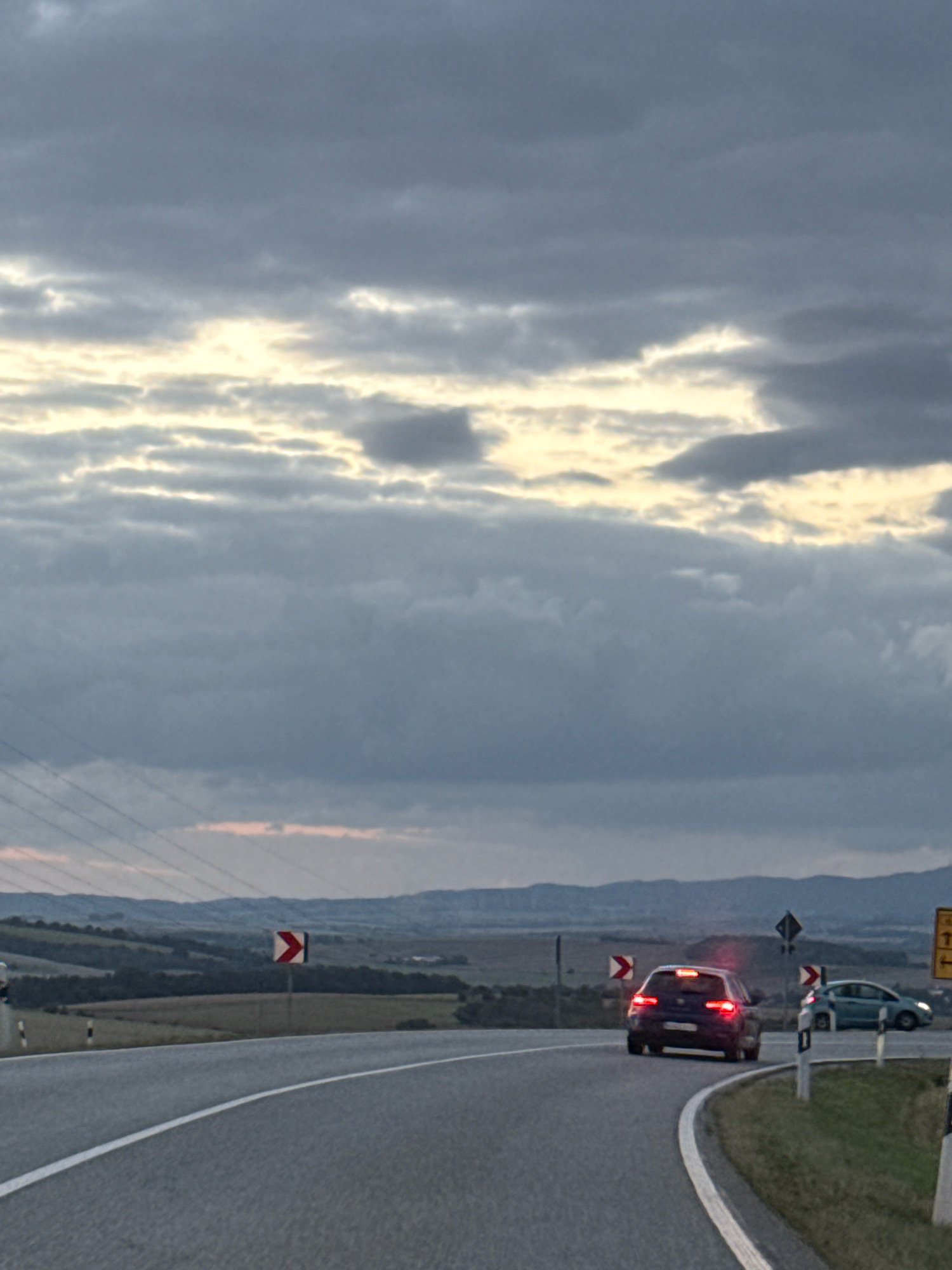 The image depicts a cloudy sky over a rural landscape with a winding road in the foreground. A dark-colored sedan is driving on the road, with its brake lights illuminated. A lighter-colored car is visible further down the road. There are traffic signs present, including a chevron sign indicating a curve in the road. The road is paved and marked with white lines. The landscape consists of rolling hills covered with vegetation and some distant mountains. There are power lines visible in the background. The overall impression is of a drive through a scenic, somewhat overcast environment. The image does not contain any information that would place it specifically in Sondershausen, Germany.