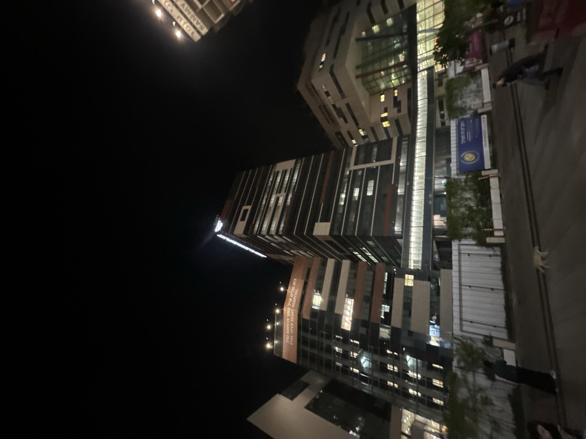 The image shows the Eyal Ofer Heart Hospital at night in Haifa, Israel. The hospital complex features modern architecture with glass facades and illuminated windows. In the foreground, there is a street with a few people walking and a dog on the pavement. There are also some trees and a white fence visible. The sky is dark, indicating it is nighttime. One of the buildings has signage that reads "Eyal Ofer Heart Hospital" in English.