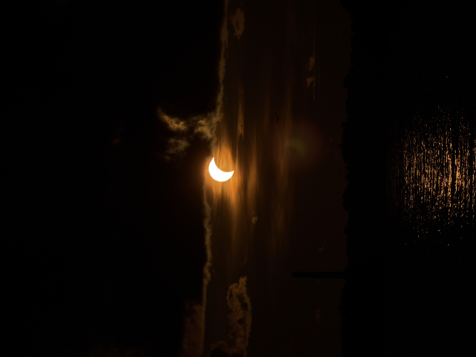 The image shows a partial solar eclipse occurring over the Daugava River in Riga, Latvia. The sun, partially obscured by the moon, casts an orange glow through wispy clouds. Silhouetted against the sky are a few distant birds and a tall, slender tower, possibly part of an industrial structure or a landmark, on the city's skyline. The lower portion of the image captures the dark, rippling surface of the river, reflecting the dim light of the eclipsed sun. The overall atmosphere is somber and dramatic.