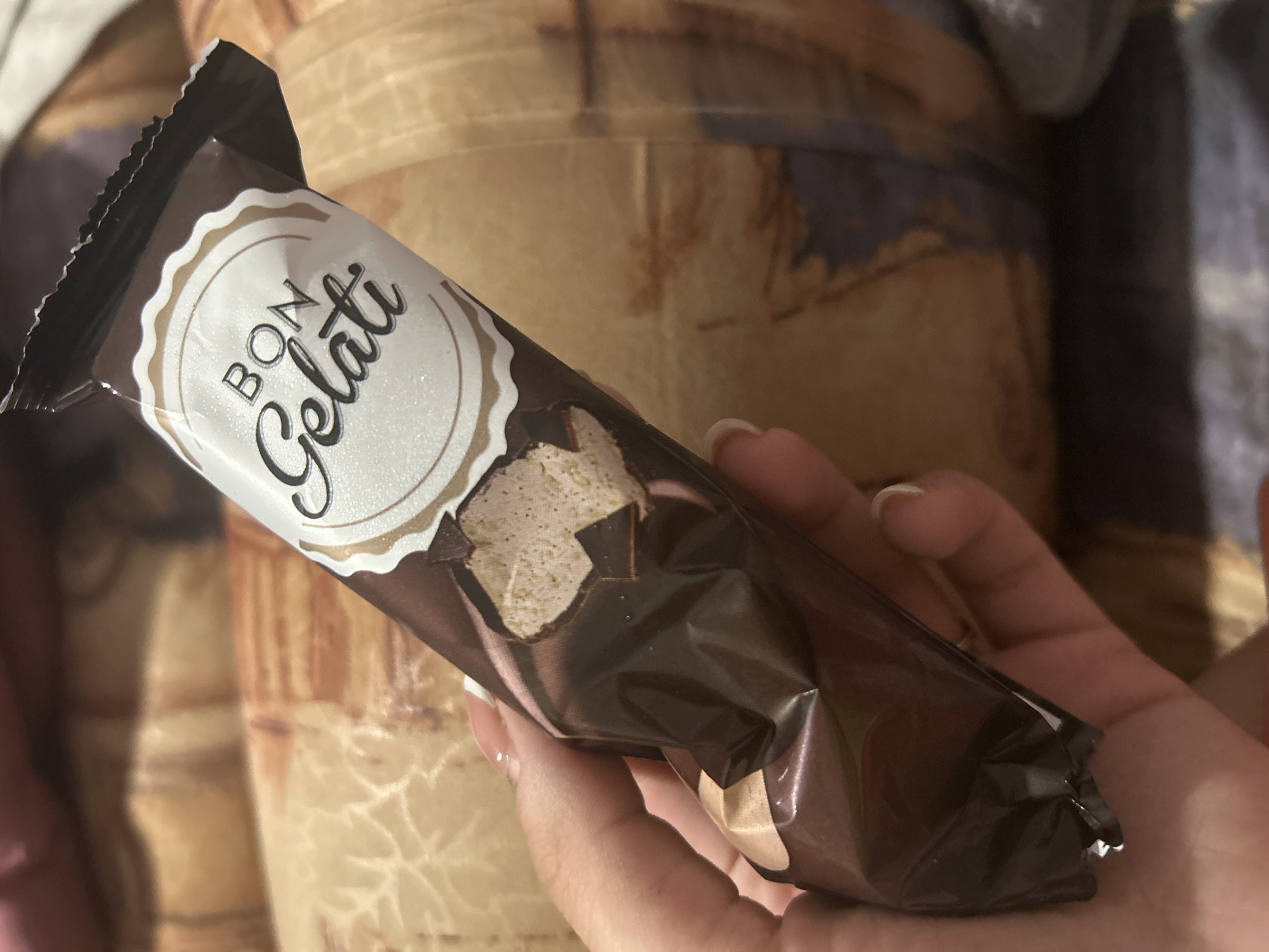 A hand holds a wrapped "Bon Gelati" ice cream bar. The wrapper is predominantly brown, with a circular white label that reads "Bon Gelati" in stylized text. A cut-out section on the wrapper reveals the ice cream inside. The background shows a patterned, presumably upholstered surface. The location in Plovdiv, Bulgaria cannot be determined from the image.