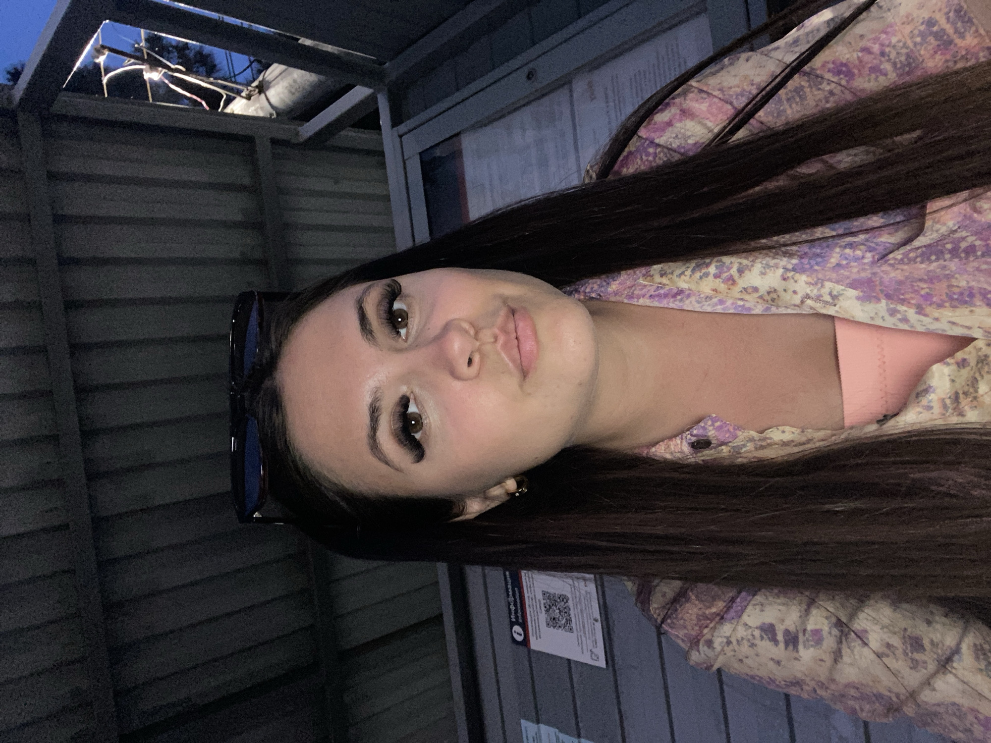 A young woman with long, dark hair is pictured outdoors at what appears to be a bus stop shelter. She is wearing a light pink top and a patterned shirt with purple and yellow floral designs. Sunglasses are perched on top of her head. In the background, there is a grey corrugated wall, a display case with information, and a tree with some wiring visible against the twilight sky. A QR code is visible on a notice posted on the wall. The image is lit by artificial light sources, contributing to the overall low-light atmosphere.