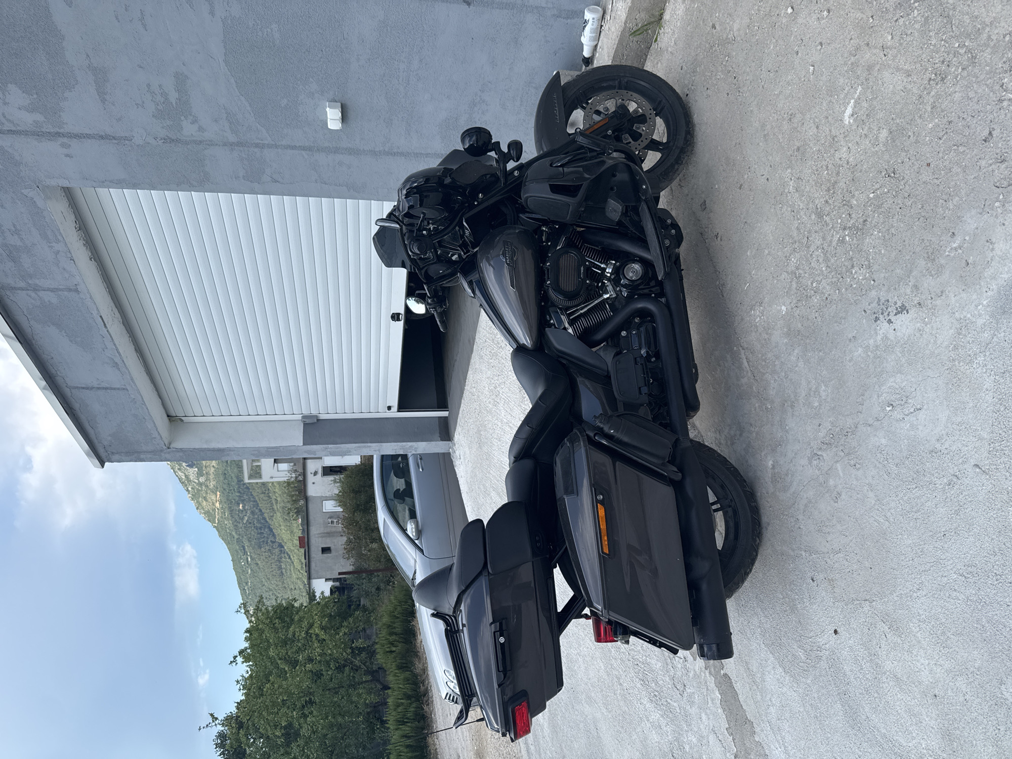 A dark grey Harley-Davidson motorcycle is parked on a concrete surface in front of a grey building with a large, white, ribbed garage door. The building is located in Radanovići, Montenegro. To the left of the motorcycle, a silver sedan is parked behind it. In the background, there are green trees and a hilly, mountainous landscape with scattered buildings. The sky is cloudy with patches of blue. A white bottle is lying on its side on the ground to the right of the motorcycle.