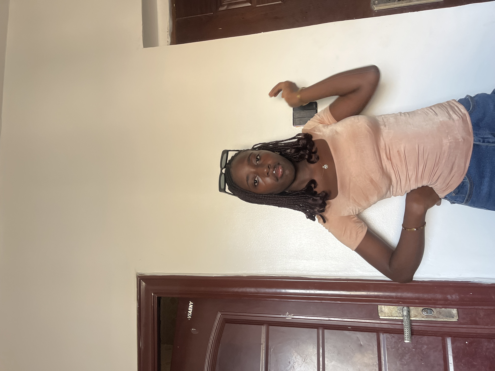 A young woman with braided hair and dark sunglasses is posing indoors against a light-colored wall, likely in Abuja, Nigeria. She is wearing a peach-colored top and blue jeans. A dark brown door frame is visible at the bottom of the image. A small black object, possibly a phone or wallet, is present on the wall next to her hand. The environment suggests an indoor space, possibly a home or apartment.