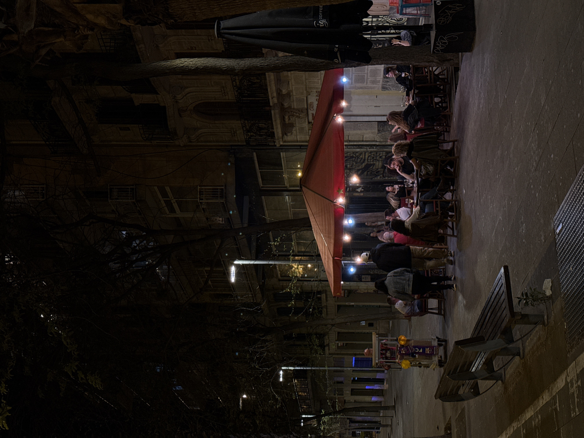 The image shows a street scene in Barcelona, Spain, at night. A group of people are seated at outdoor tables under a red awning decorated with string lights. Some are eating and drinking, while others are engaged in conversation. An umbrella with the "fig Coffee & tea" logo is visible near the front of the building. The buildings along the street are lit by streetlights, and a bare tree is partially visible on the left side of the image. A park bench sits vacant at the edge of the street on the right, next to a drain.