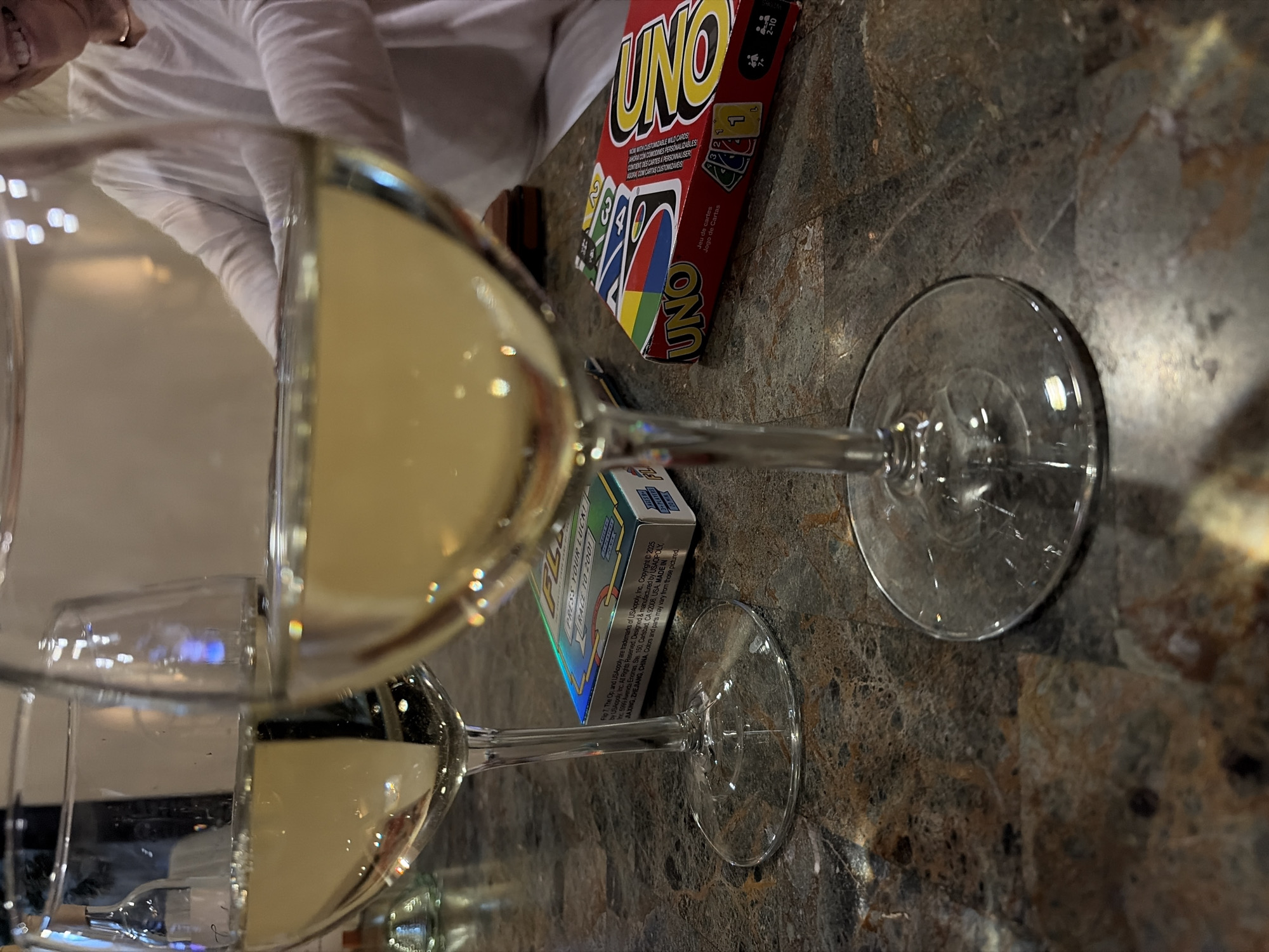 A tabletop scene, photographed at a close-up angle, shows two wine glasses filled with a light-colored beverage, likely white wine. Next to the wine glasses, a box of UNO cards and another box of "FL FLIP" game cards can be seen. In the background, a person wearing a white shirt is partially visible. The table has a dark, patterned surface resembling marble or granite. It can be inferred that this photograph was taken in the city of Trophy Club, United States, from the OCR text of the card.