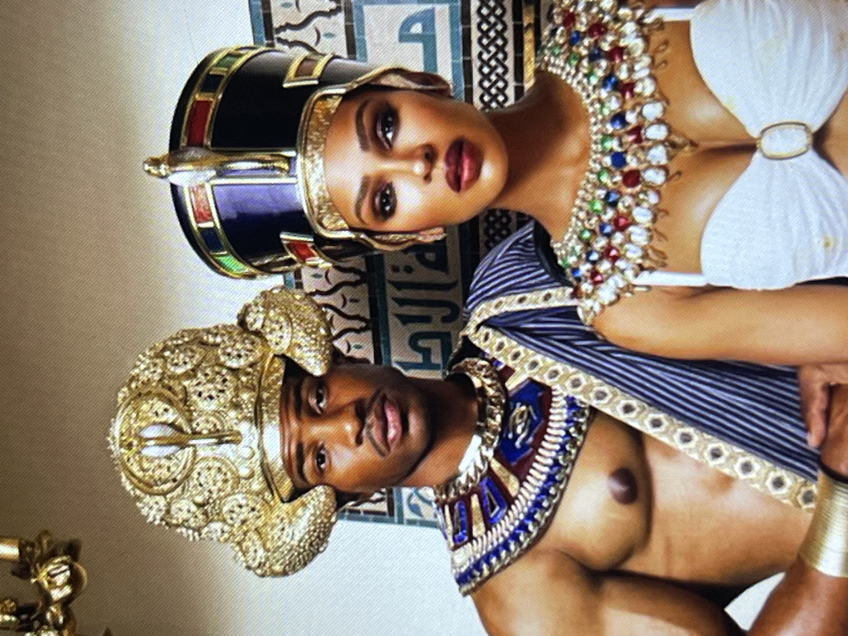 The image, captured in Muchiya, Nigeria, presents two individuals, an adult male and an adult female, posed in elaborate attire evocative of Ancient Egypt. The male, positioned on the left, has a dark complexion and a muscular build, with a slight mustache and soul patch. He wears a large, ornate golden headdress resembling a pharaoh's crown, featuring a prominent uraeus cobra at the front. A broad, multi-layered collar necklace in shades of blue, red, and gold is worn around his neck, and a blue and gold striped sash is draped over his left shoulder. His torso is bare. The female, positioned on the right, also with a dark complexion, wears a dark blue crown with gold bands and a uraeus cobra. She has a large, ornate collar necklace encrusted with multiple colorful gemstones (red, green, blue, clear) and gold drops, covering her upper chest. She wears a white, strapless top with a central gold ring detail. Both individuals maintain a direct gaze towards the viewer. The background consists of a wall adorned with decorative blue and white tiles, featuring geometric patterns and what appears to be stylized Arabic script. A segment of a golden chandelier is visible in the upper left corner.