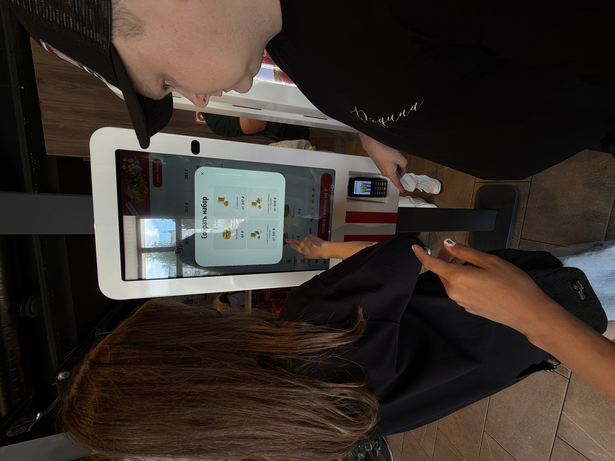 The image shows two people, a woman with long brown hair wearing a black t-shirt and a man wearing a black t-shirt and a black baseball cap, standing in front of a self-service ordering kiosk. The kiosk screen displays a menu with food items and prices in Russian. The woman is reaching out to touch the screen, and the man is looking at the screen. A small mobile phone is attached to the kiosk. The environment appears to be an indoor fast-food restaurant, possibly a McDonald's, indicated by the kiosk design and the menu items visible on the screen. The floor is tiled. The location is identified as Chaikovsky, Russia.