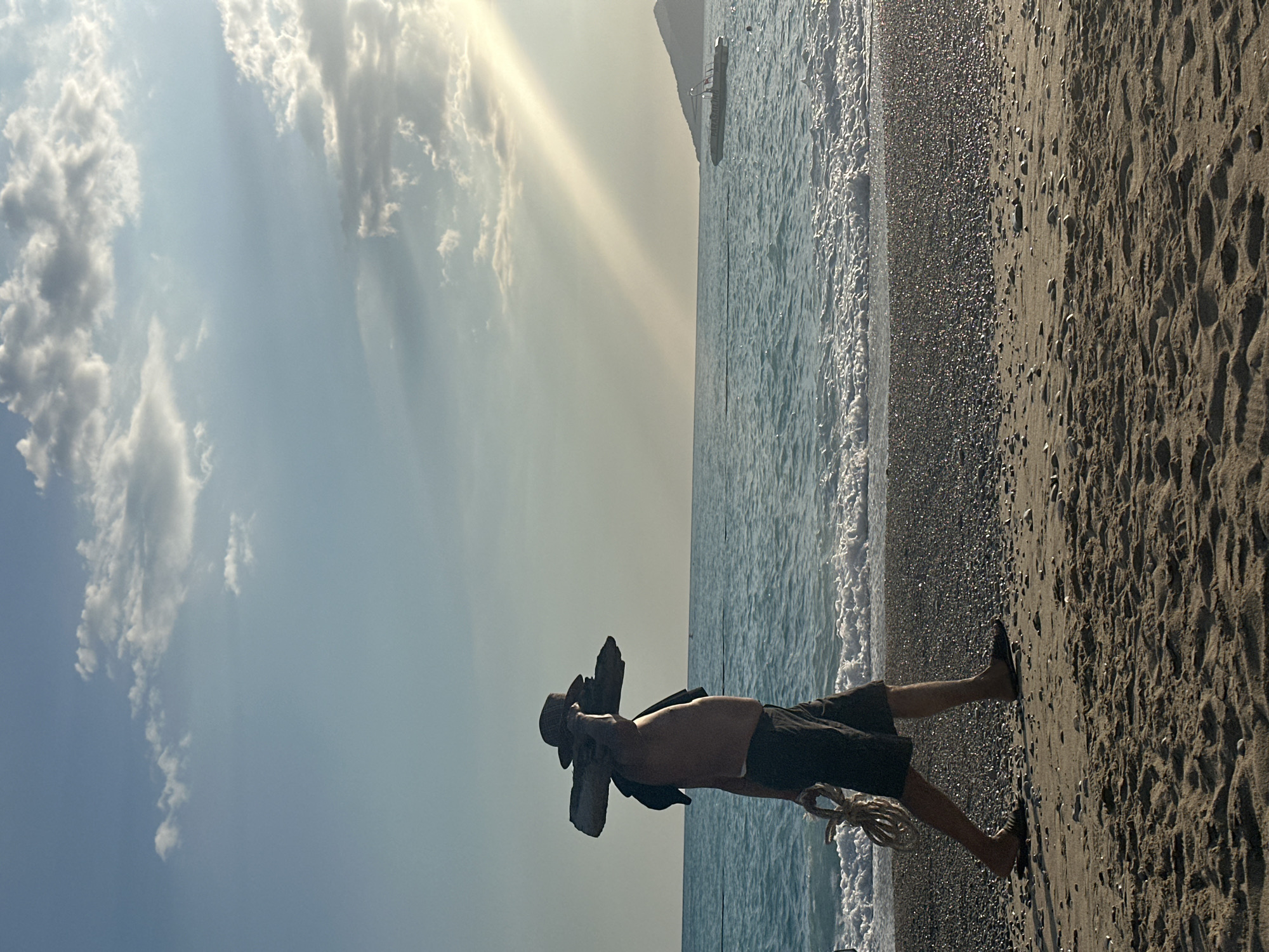 A horizontal, long-shot photo shows a beach in Fethiye, Turkey, with a shirtless man holding a large piece of driftwood. The background features a bright sky with sun rays through clouds, and a calm ocean with gentle waves lapping the shore. The beach transitions from the wet sand and pebbles near the water to drier sand further inland. The man stands on the beach, facing the water, with the driftwood held in front of him. He wears dark shorts and appears to have a dark jacket over his shoulder. A rope hangs down from the driftwood. The lighting suggests it is either sunrise or sunset. A small boat rests in the water in the distance.