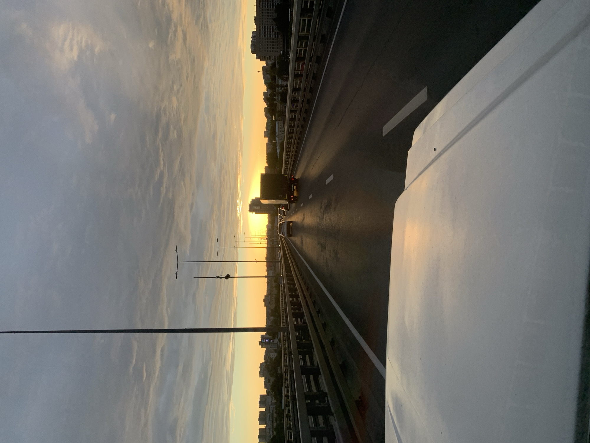 The media file depicts a view from a vehicle driving on a highway bridge over a city during sunset. The sky is filled with dramatic clouds illuminated by the warm, golden light of the setting sun, creating a sense of awe. In the distance, the silhouette of a city skyline with tall buildings emerges from the horizon. The highway is wet, suggesting recent rain, and traffic, including a truck and several cars, is visible moving along the lanes. The presence of streetlights indicates it is an urban environment. The image is taken from the perspective of someone inside or on top of a white vehicle, the hood of which is prominently featured in the foreground on the right side of the frame. The overall mood is serene and reflective, capturing the beauty of a sunset over an urban landscape. The time of day is clearly sunset, and the weather appears partly cloudy with a possibility of recent rainfall. The location context points to Nizhny Novgorod, Russia. There is no visible text in the media file.