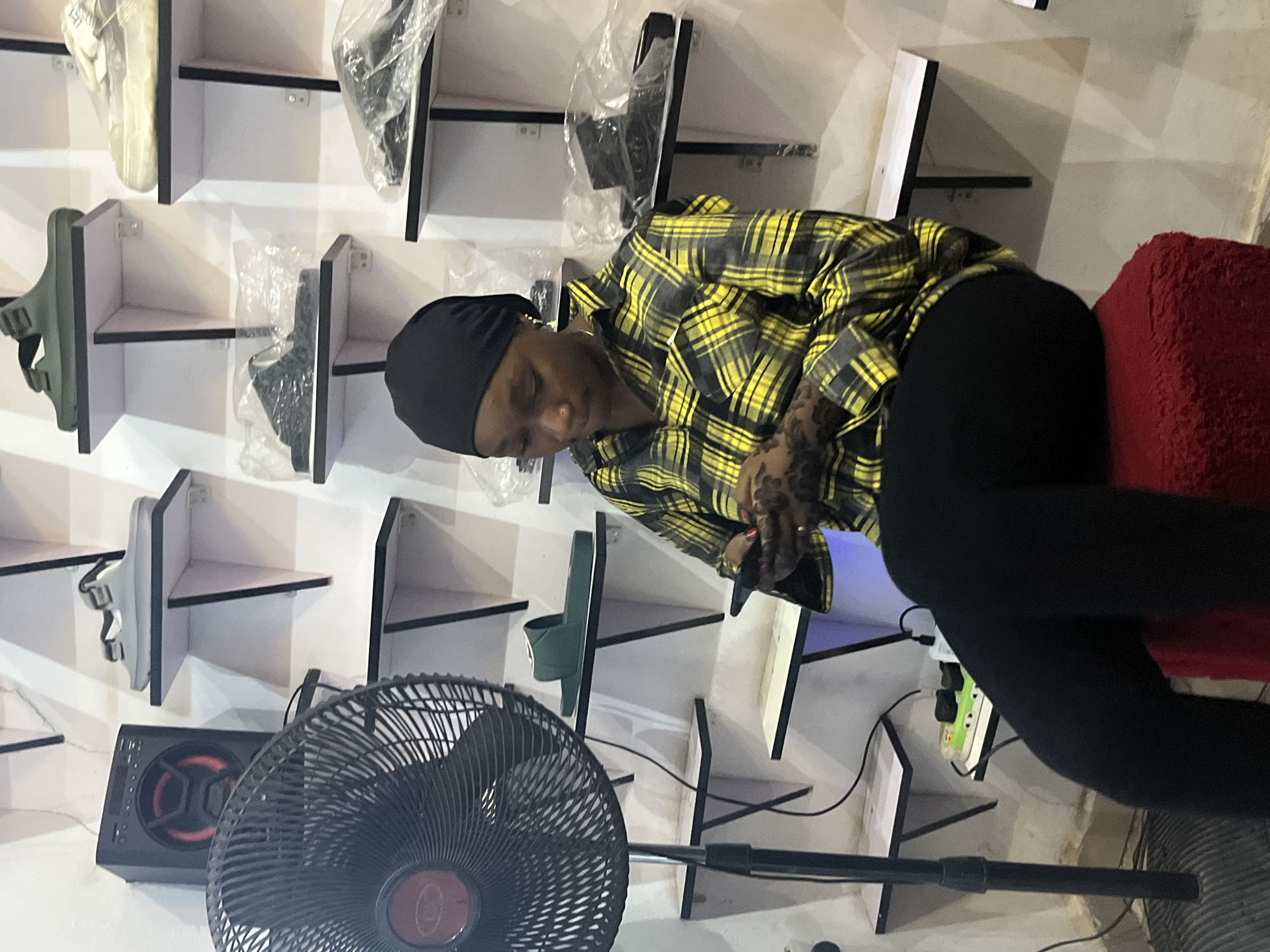 In a store that appears to be in Jos, Nigeria, a young woman wearing a black headscarf and a yellow and black plaid shirt is seated on a red stool, looking down at her mobile phone. She has intricate henna designs on her left forearm. Behind her, shelves are stacked with various types of footwear, including sandals and sneakers. To her left, a black standing fan is positioned. The overall environment suggests a retail setting for clothing or accessories.