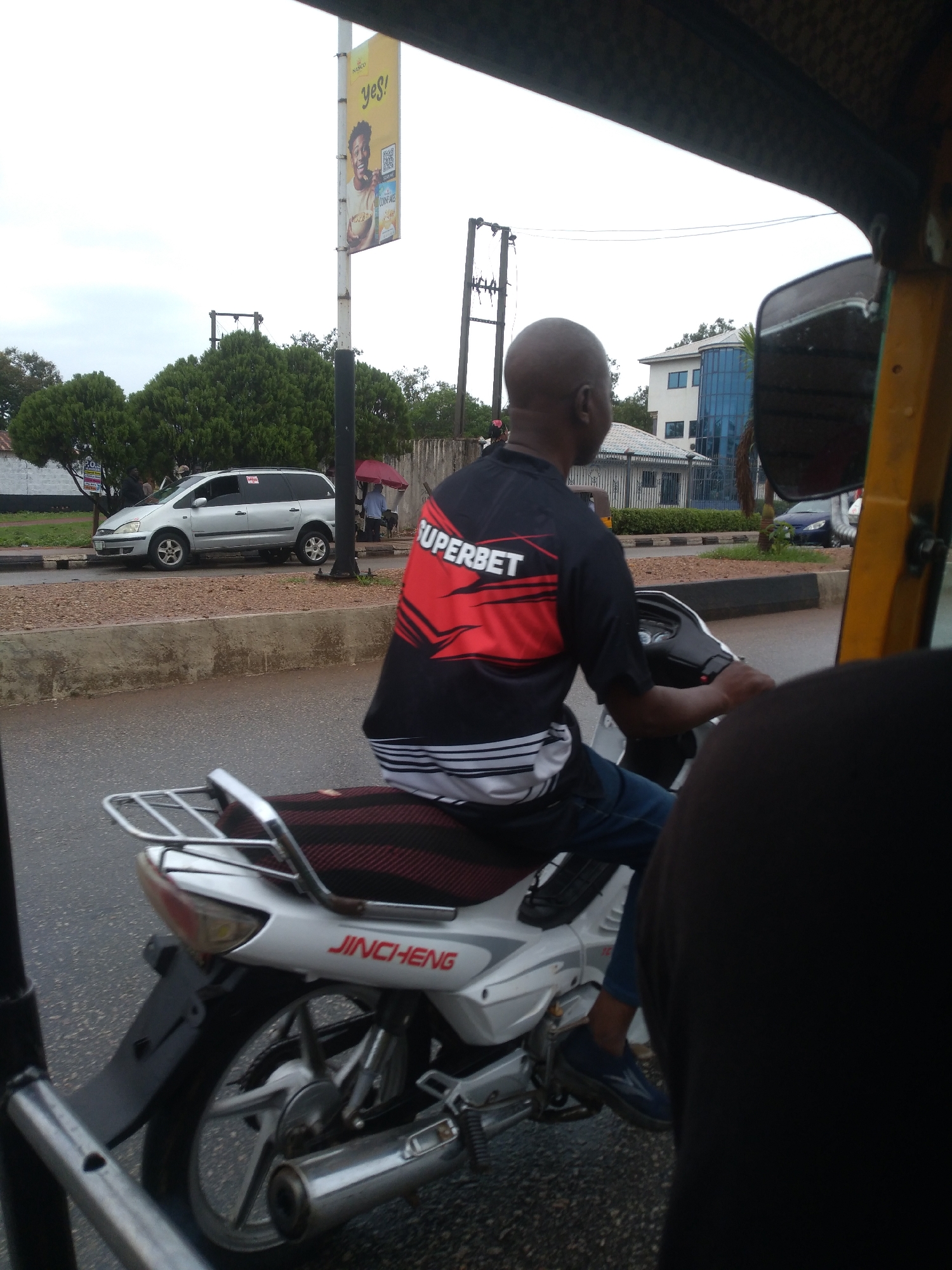 A street-level photograph captured from within a vehicle, likely a tricycle or similar open-sided transport, shows a man on a white Jincheng motorcycle in the foreground, viewed from behind. He wears a black and red "SUPERBET" branded shirt and blue jeans. The road appears wet, suggesting recent rain.
In the midground, a silver minivan, possibly a Ford Galaxy, is parked or driving on the left side of the road, with people standing nearby, one under a red umbrella. Beyond the road, there's a concrete median strip with some greenery, and further back, various buildings, including one with a distinctive blue glass facade.
On the right side of the image, the frame includes a portion of the photographer's vehicle and its rearview mirror. Overhead, an overcast sky dominates, indicating daytime. A tall pole stands prominently in the midground, bearing a yellow advertisement that reads "Yes!" and features a person holding a bowl of "Nasco" branded "CORN FLAKES" along with a QR code. The overall scene depicts a busy, possibly urban or semi-urban environment.