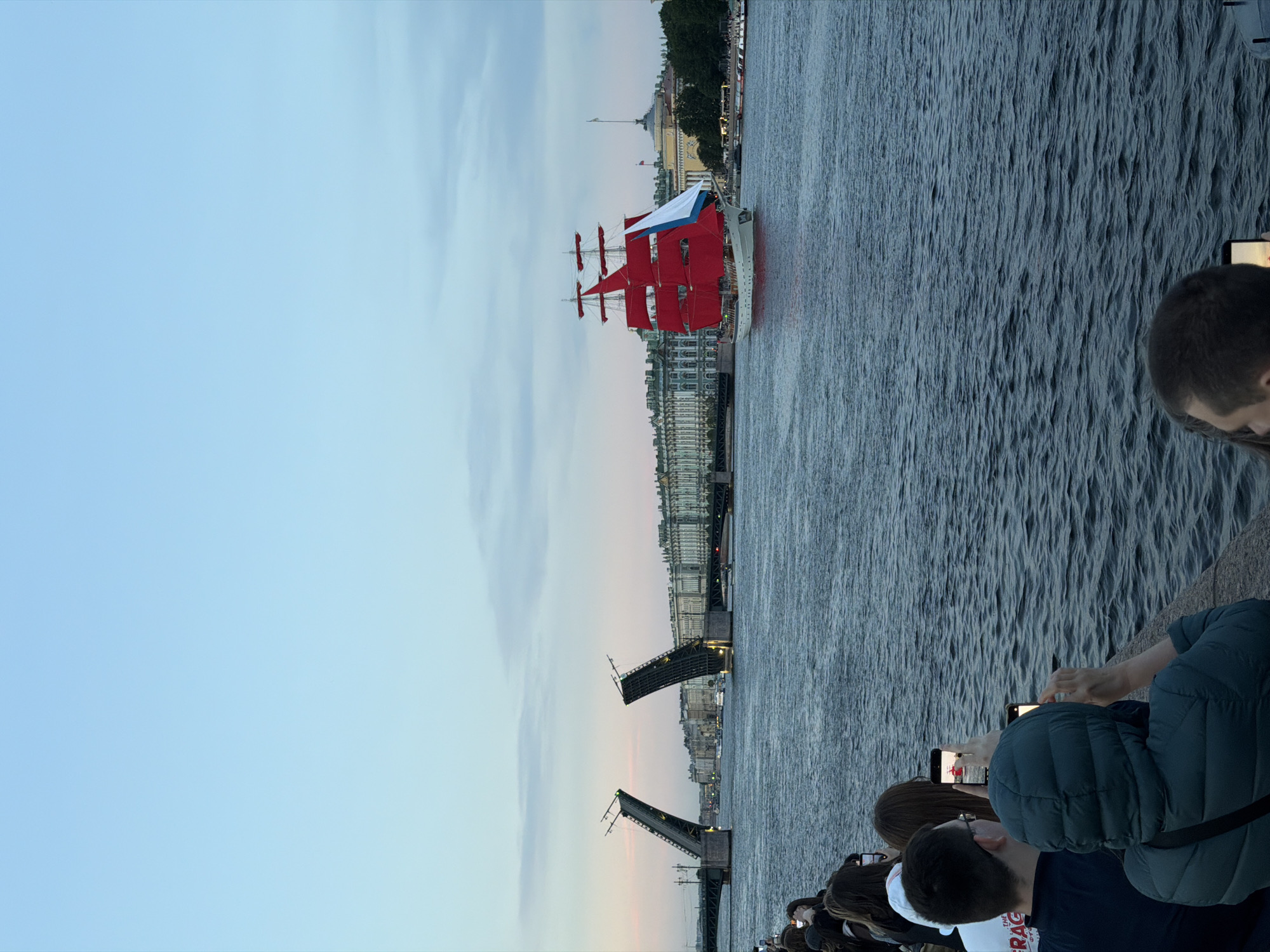 The image captures a scene along the Neva River in Saint Petersburg, Russia, at dusk. In the background, the Palace Bridge is raised, its two halves angled upwards to allow passage for vessels. Along the embankment, a grand row of classical buildings lines the horizon. The focal point of the scene is a magnificent three-masted sailing ship with vibrant red sails, cruising on the river. A portion of its sails are a crisp white, and a blue flag is visible. In the foreground, a group of people are gathered along the riverbank, many holding up their mobile phones to capture the spectacle. Their backs are mostly to the camera, indicating their attention is directed towards the ship and the bridge. The water is a deep blue, reflecting the muted colors of the sky, which displays soft hues of pink and orange from the setting sun.