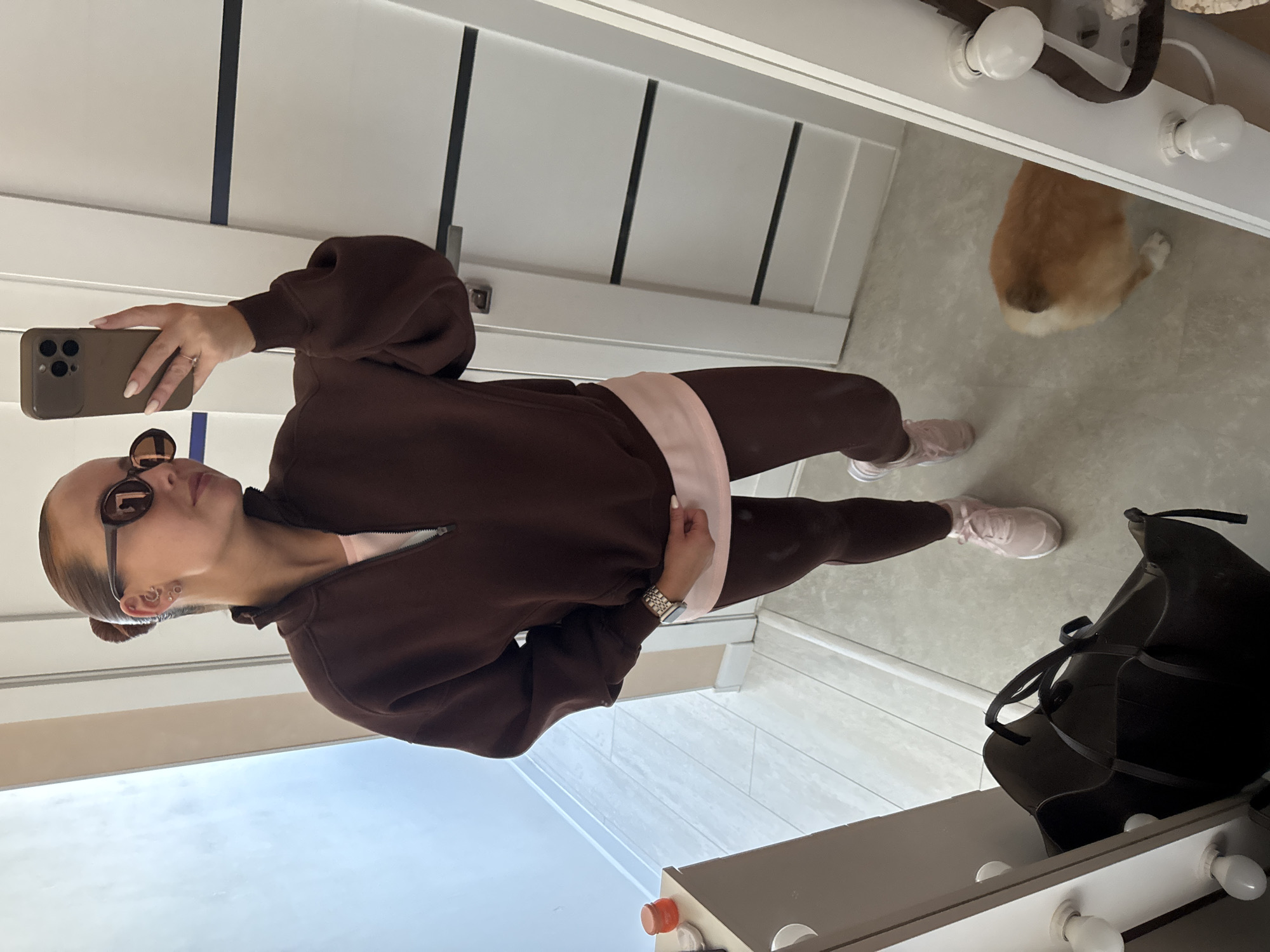 A woman is taking a selfie in a room with a mirror. The woman is wearing sunglasses and a brown tracksuit with a pink shirt underneath. She is holding a smartphone with her right hand, and her left hand is on her hip. In the background, there is a white door with black stripes, a black purse, and a ginger cat. The location cannot be determined as Chaikovsky, Russia, based on visual information.