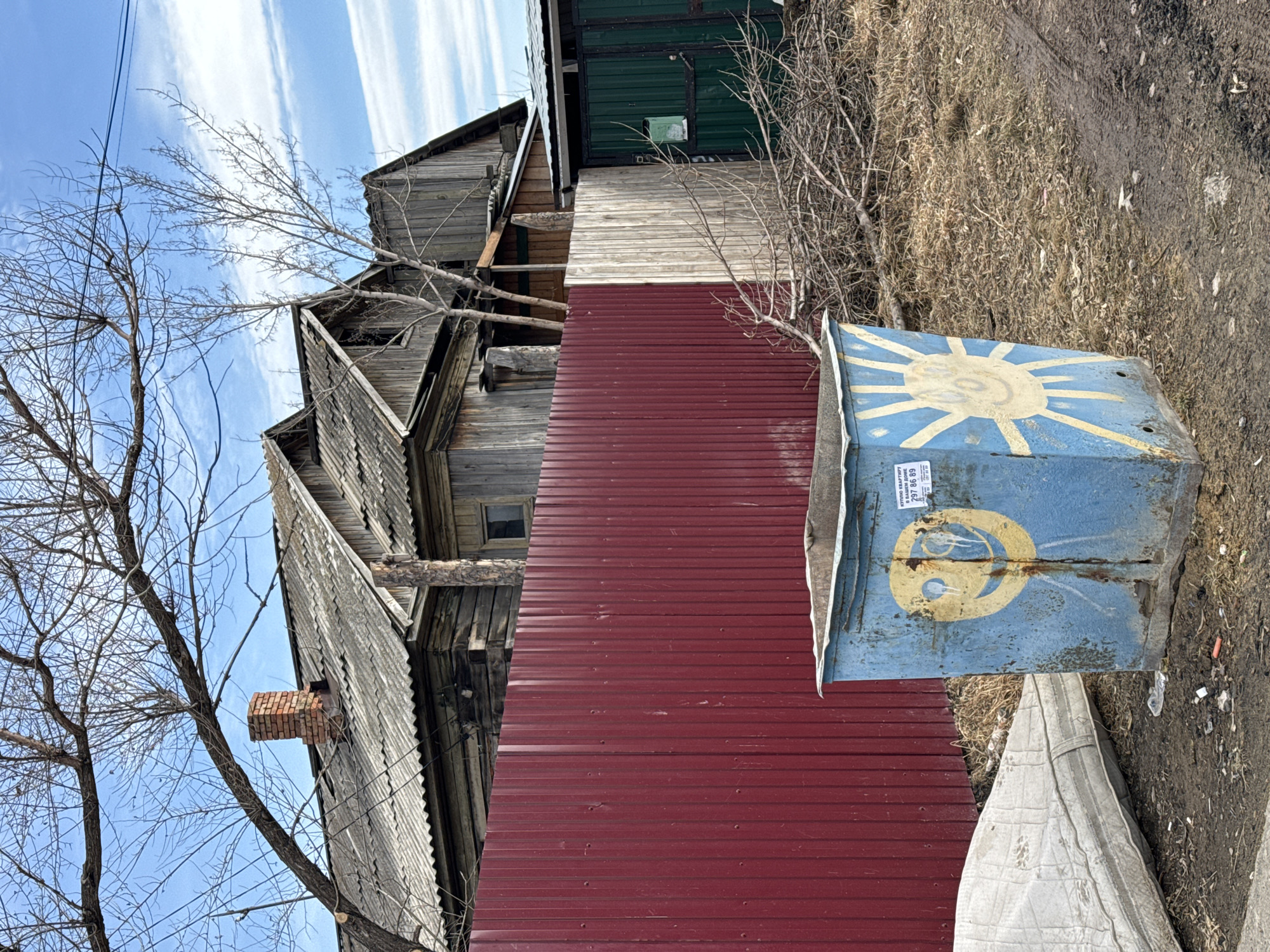The image shows an old, three-story wooden house with peeling grey paint next to a red corrugated metal fence. A bare tree stands in front of the house. Adjacent to the fence is a blue, weathered trash container with a yellow sun painted on the lid and a partial yellow crescent moon design painted on the side. A faded sticker with the numbers "297 86 89" is on the side of the container. A soiled, light-colored mattress is partially visible near the base of the container. The ground appears to be a mix of dirt, sparse vegetation, and debris. The sky is blue with some streaky clouds. Based on the architectural style and the Cyrillic numerals on the container's sticker, it's likely that this image depicts a scene in Russia. Further context is that this image shows a location near the city of Krasnoyarsk, Russia.