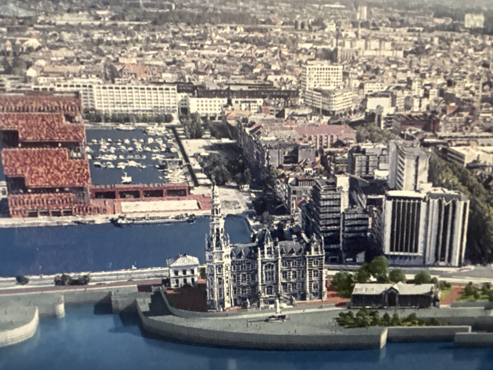 This is a vertical aerial view of a city with a prominent waterway on the left side. The waterway is bordered by a curved embankment on the far left, with a sidewalk or promenade running alongside it. Adjacent to the waterway are several buildings, including a large complex of grey multi-story structures with numerous windows and architectural details, possibly a historical district or a well-preserved older part of the city.
Further inland, the cityscape transitions to a dense urban environment with a mix of building styles and heights. There are numerous brick buildings, some with red or reddish-brown facades, and modern white or grey structures with grid-like window patterns. A section of the waterway appears to be a harbor or marina, with many small boats docked. Beyond this, the buildings become more densely packed and extend as far as the eye can see, indicating a large urban area. The image is somewhat blurry and has a grainy quality, suggesting it might be a photograph taken from a distance or a scanned print. The lighting suggests it is daytime, with shadows visible on some buildings. There are no people or vehicles clearly visible in the image.