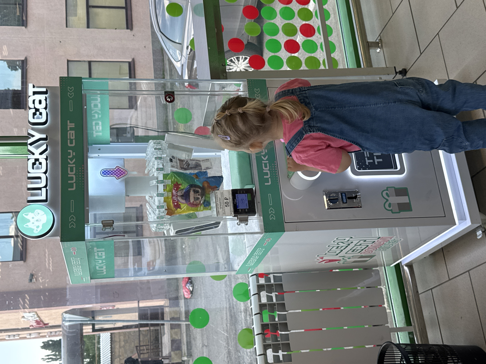 The image shows a young girl in a pink shirt and denim overalls playing with a "Lucky Cat" claw machine. The machine is white and green with colorful circles on the glass. There are prizes inside the machine, including a bag of "Labubu" toys. The girl is looking intently at the machine, her hands near the controls. The machine has text in Russian, including "СИМУЛЯТОР ЛОРКОСТИ" ("Simulator of luck") and "50 P" (likely the price per play). The setting appears to be outdoors, possibly in a shopping area or entertainment venue, given the reflection of buildings and trees in the glass of the machine. The lighting suggests it is daytime. The girl's focus and the presence of a game suggest she is engaged in playing and enjoying herself. The machine itself is a prominent object, decorated with the "Lucky Cat" logo and branding.