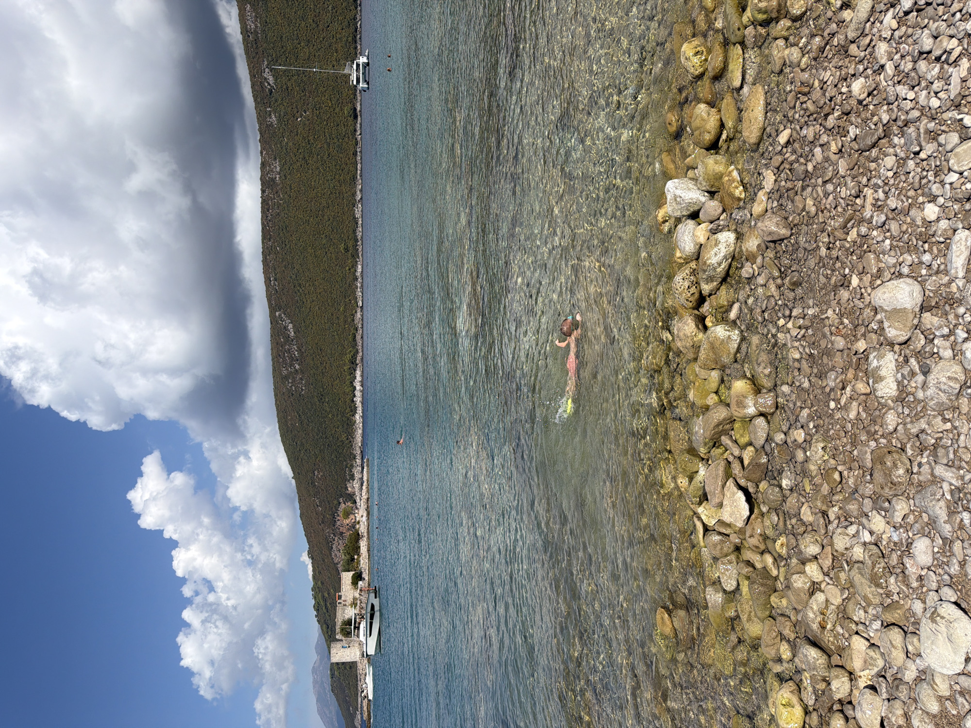 A person is swimming in the clear, shallow water near a pebbled shore. The swimmer is wearing a pink swimsuit and a snorkel mask. The water is transparent, allowing the rocky bottom to be visible. In the background, there are boats anchored in the sea, and a lush green hill slopes down to the water's edge. A stone tower and some buildings are visible on the shore in the distance. The sky is blue with scattered white clouds. The scene appears to be a coastal area in Luštica, Montenegro.