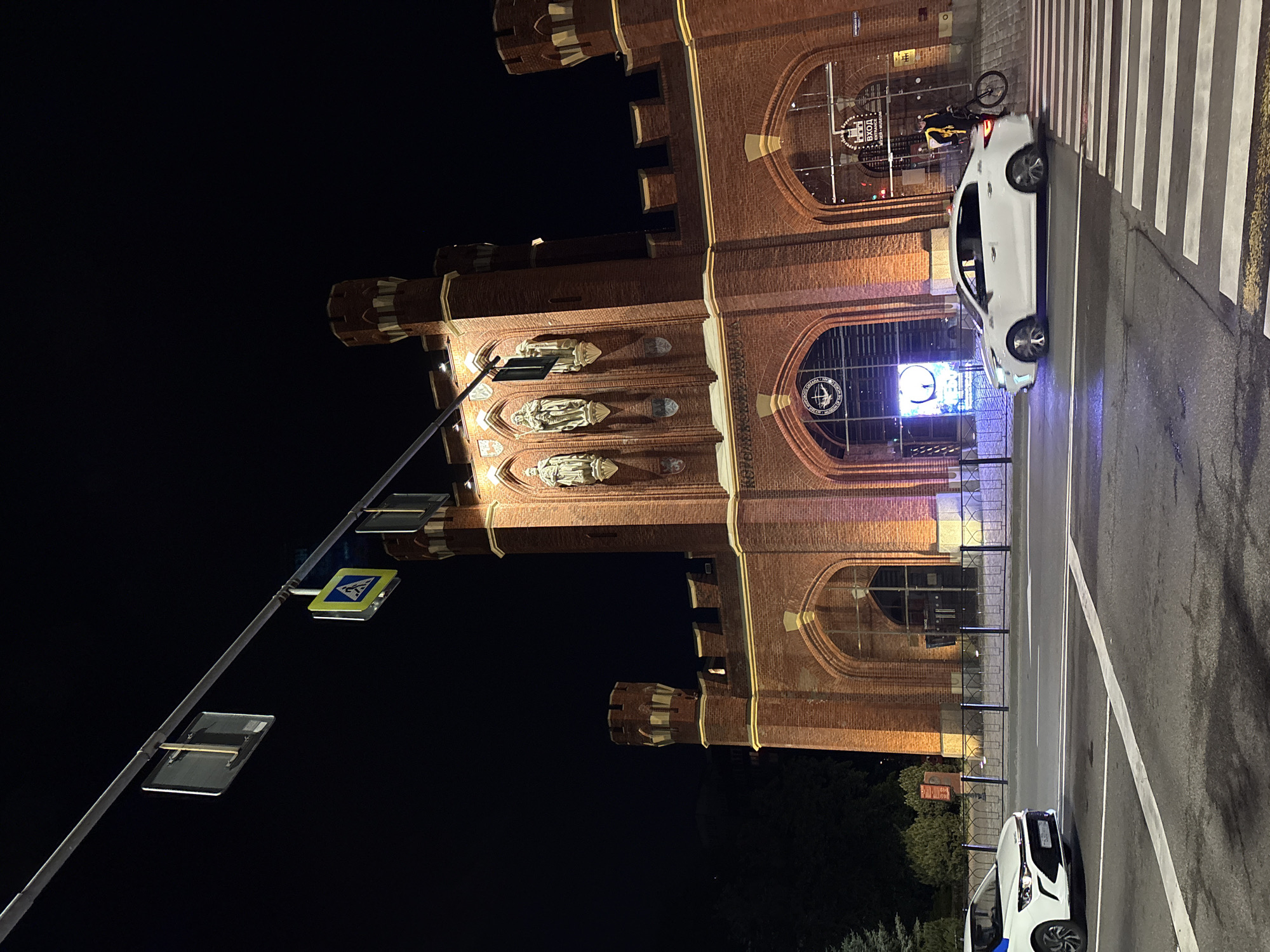 The media file captures a night-time street scene in Kaliningrad, Russia, showcasing a large, illuminated brick building with a historical or Neo-Gothic architectural style. The building features intricate brickwork, multiple arched windows and entrances, battlements, and statues within niches on its facade. Warm artificial lighting highlights the building's details against the dark night sky.
In front of the building, a paved road with a clearly marked pedestrian crossing (zebra crossing) is visible. Two white cars are present; one is parked or stopped at the pedestrian crossing, partially obscuring a bicycle with an indistinct person. The other white car is partially visible in the bottom right. A tall lamppost or utility pole on the left features flat, rectangular elements and a yellow pedestrian crossing sign.
Notable details on the building include a large clock face glowing blue within an arched window. Another prominent arched window displays the illuminated Russian word "ВХОД" (VKHOD), meaning "ENTRANCE," along with smaller text that appears to be "РЫБНАЯ ДЕРЕВНЯ" (RYBNAYA DEREVNYA), or "FISH VILLAGE." Another window features an emblem with a nautical theme, possibly a ship's wheel or compass. The setting suggests an active urban area, likely a tourist or commercial district, given the building's prominent design and visible signage.