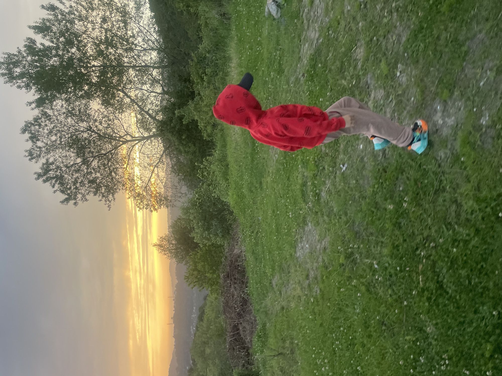 A person wearing a red hooded sweatshirt and a black baseball cap is standing on a grassy field. They are wearing tan pants and multi-colored sneakers. Behind them, a tree is silhouetted against a sunset. In the distance, there is a hazy view of a town. The town in the distance is likely Baranovka, Russia.