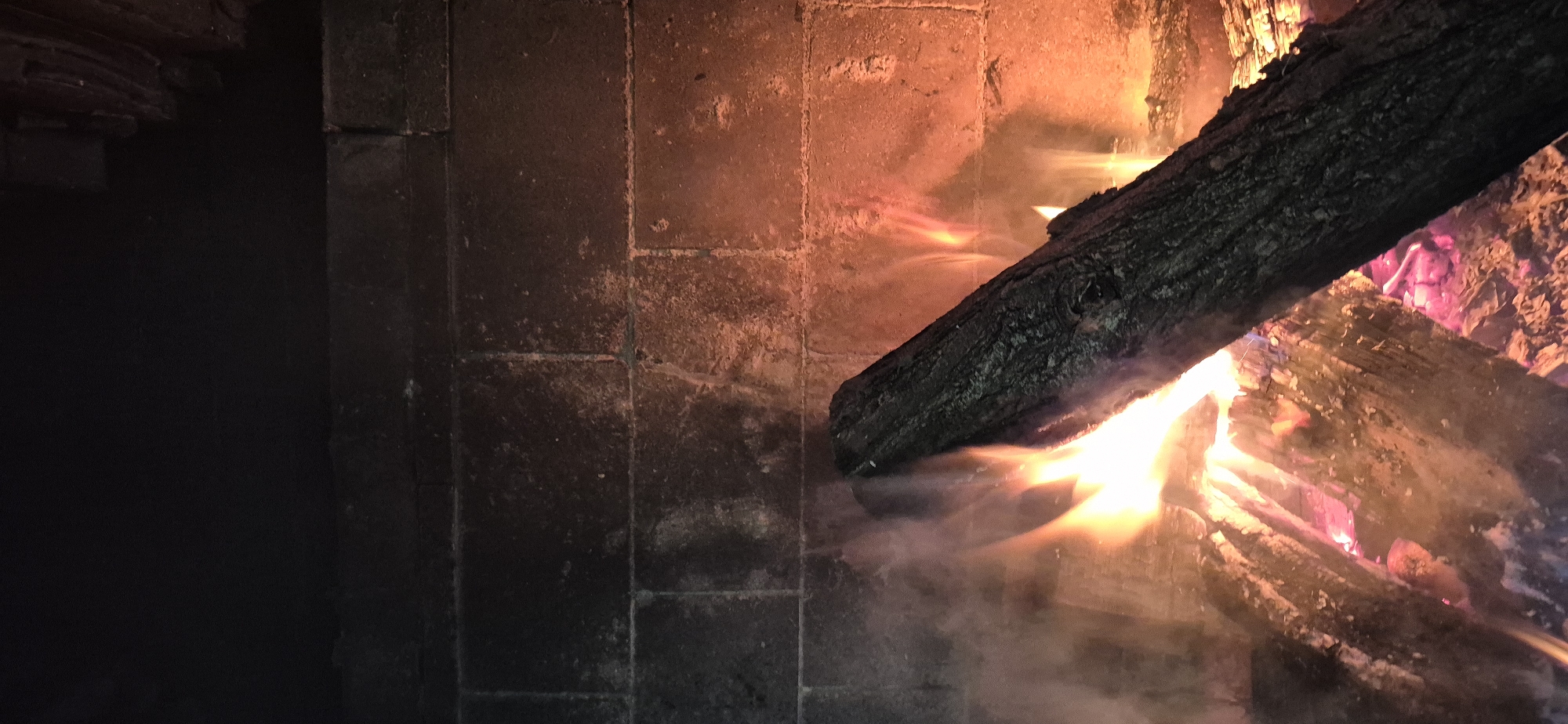 A log fire burns in a brick fireplace. The fire emits smoke and a warm orange glow that illuminates the dark bricks of the fireplace. The top of the fireplace is black and appears to be a chimney opening. The scene appears to be a domestic setting.