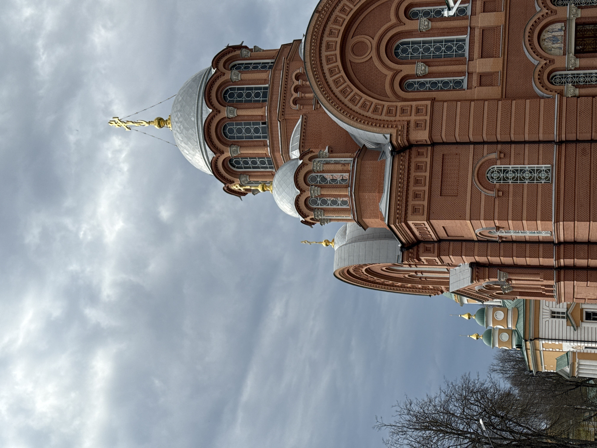 The image shows a detailed view of a large, brick Orthodox church with multiple domes and intricate architectural details. The church is located in an unspecified city, as its location is noted as Unknown, Unknown. The primary colors are brick red, silver (for the domes), and gold (for the crosses atop the domes). The sky in the background is overcast with streaks of white and gray clouds. At the bottom right of the image, portions of other buildings with yellow and white facades and green-colored domes are visible. Bare tree branches can be seen in the very bottom center of the image.