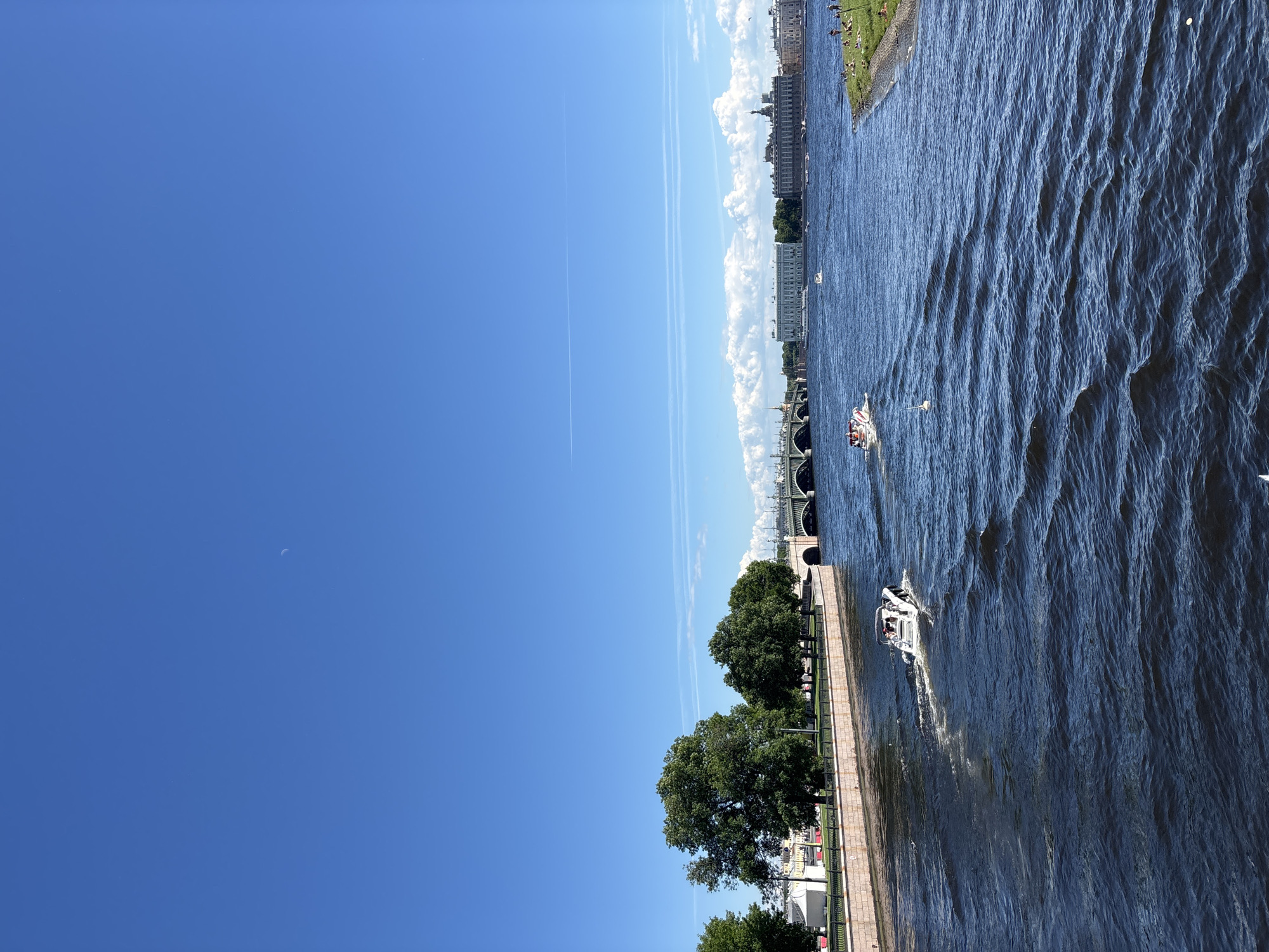 This is a wide-angle, landscape, eye-level photograph of the Neva River in Saint Petersburg, Russia, on a clear, sunny day. The sky is a vibrant blue with a faint, crescent moon visible high in the upper left quadrant. A thin, white contrail streaks across the sky.
In the foreground, the dark blue water of the Neva River is choppy with visible waves. Two small white motorboats are moving through the water. The boat closer to the camera is moving away from the viewer and to the left, leaving a white wake. The second boat, further in the distance, is also moving away from the viewer and to the left.
Along the left bank of the river is a stone embankment lined with a low, black metal fence. Behind the fence are several mature, lush green trees. A light-colored building is partially visible behind the trees on the far left.
In the midground, spanning the river, is a large, green arched bridge with multiple spans. Beyond the bridge, the cityscape of Saint Petersburg is visible. Prominent buildings with light-colored facades and architectural details are lined up along the riverbank. Some smaller boats are dotted on the water in the distance.
On the right bank of the river, a grassy area slopes down to the water. Several people are visible on the grass, some lying down, appearing to be sunbathing or relaxing.
The overall scene depicts a lively urban riverfront on a beautiful summer day.