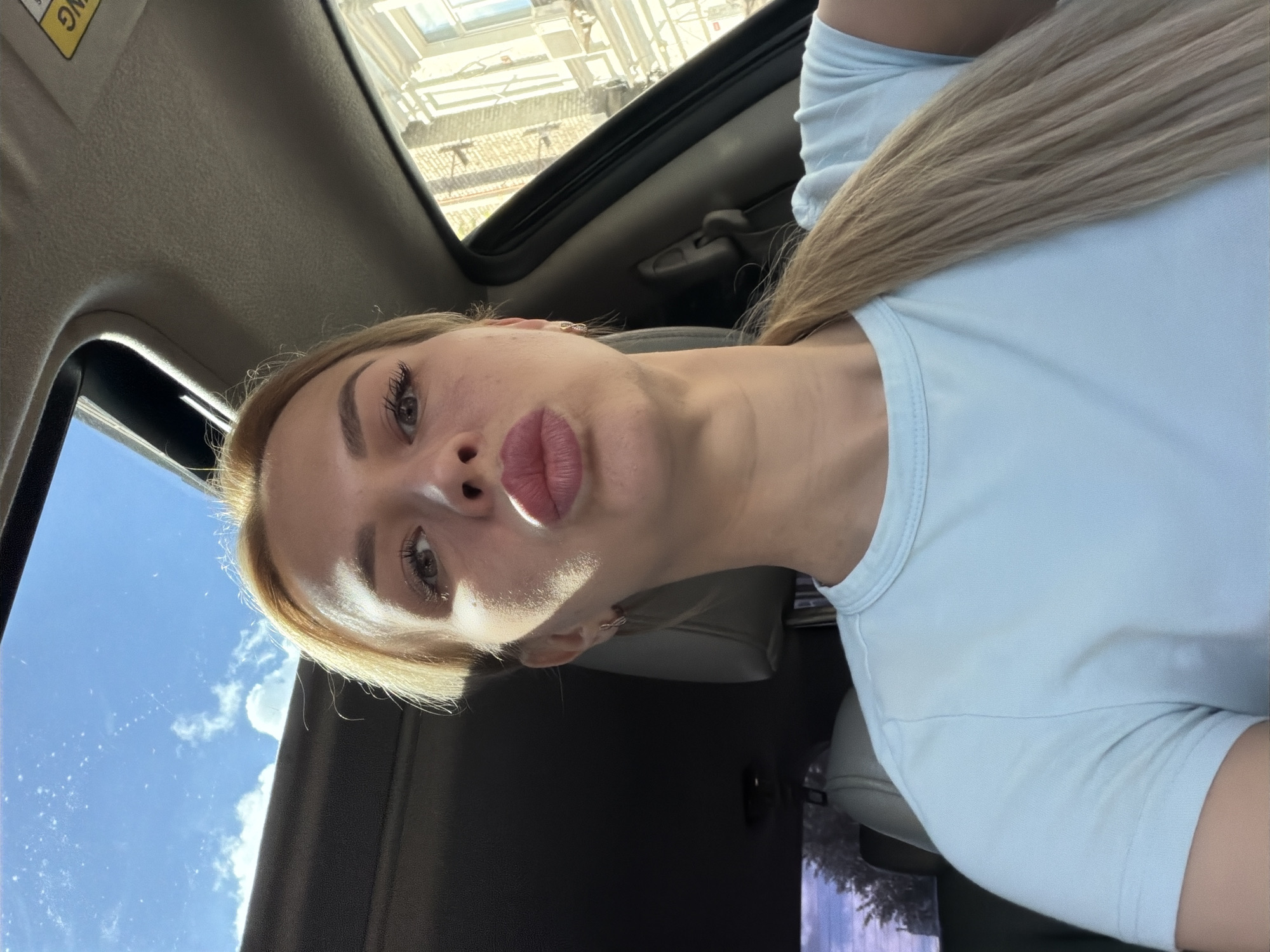 A woman with blonde hair and a light blue t-shirt is inside a car, looking at the camera. She is puckering her lips. The car has a sunroof open, revealing a blue sky with clouds. Buildings are visible outside the car window. The scene is in Kyiv, Ukraine.