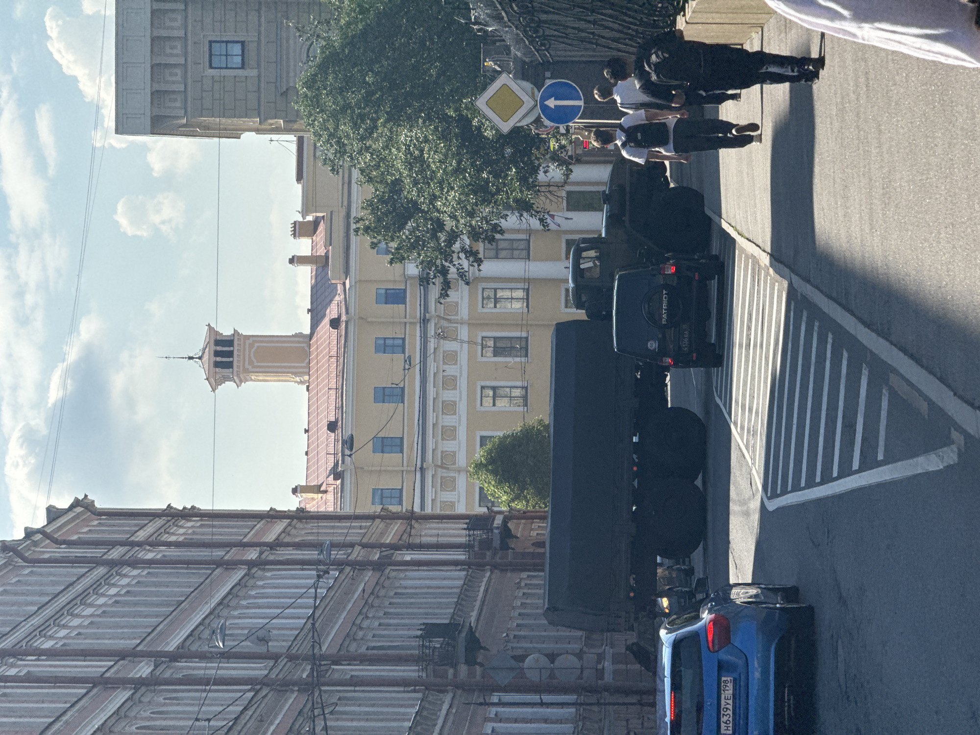On a sunny day in Saint Petersburg, Russia, a military truck with a large dark tanker trailer is parked on a city street, likely near the Peter and Paul Fortress due to the architectural style of the surrounding buildings. The truck is positioned behind a blue Mercedes-Benz A-Class. Two young men wearing casual clothing and backpacks walk down the sidewalk, moving away from the viewer. The sidewalk is bordered by a dark metal fence. In the background, a yellow building with a tower stands out against a partly cloudy sky. The street is paved, and there are traffic signs, including a diamond-shaped yellow sign with a black border and a blue circular sign with a white arrow. The buildings on either side of the street are multi-story with classical architectural features.