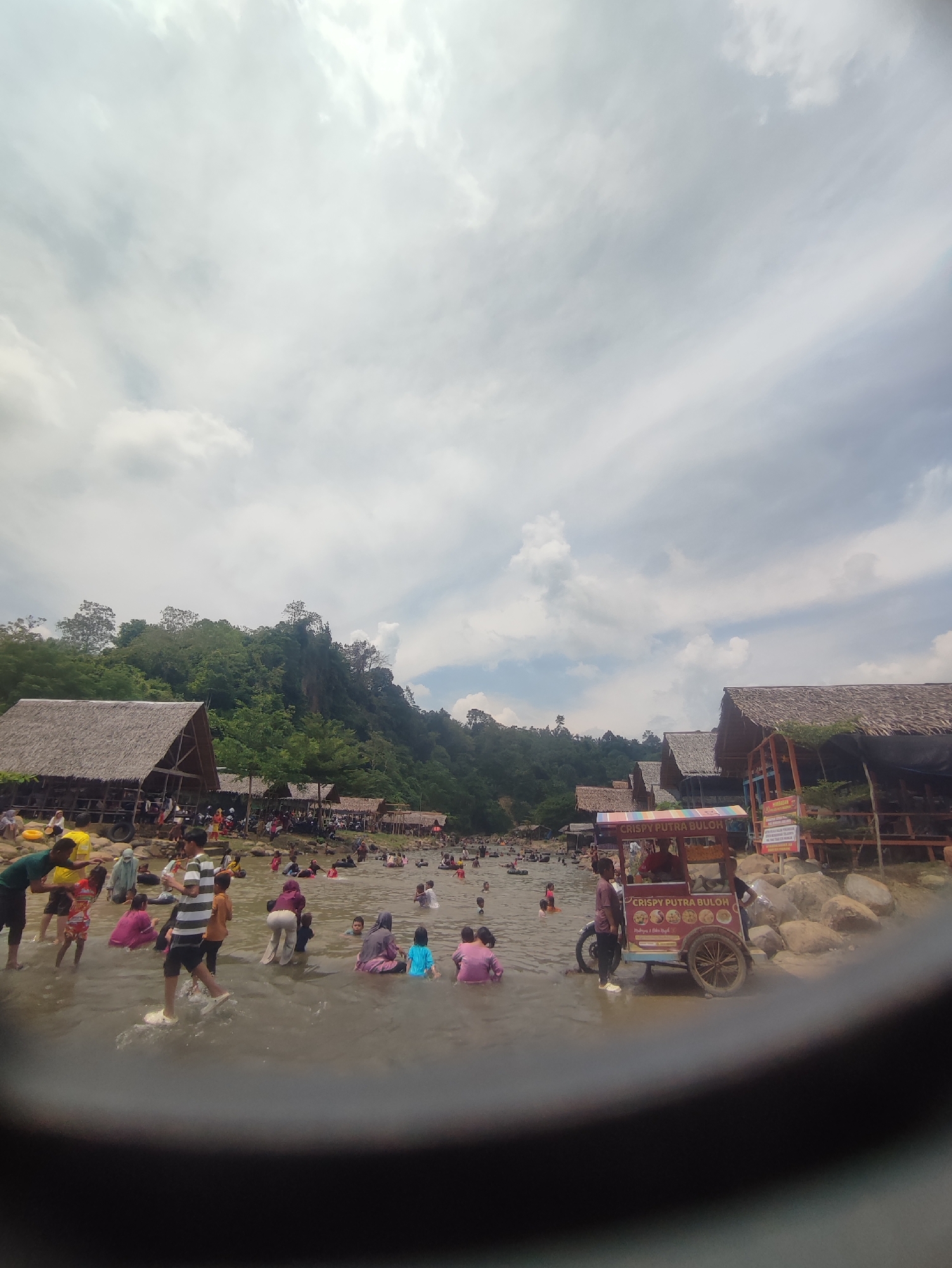 A group of people are enjoying a day at the river in Pantonrayeuk Satu, Indonesia. The river is shallow and many people are wading in the water. There are several huts with thatched roofs on the riverbank, and a food cart selling crispy buloh. The sky is cloudy, but the sun is shining through in some areas. The river is surrounded by lush green trees and a hilly landscape.