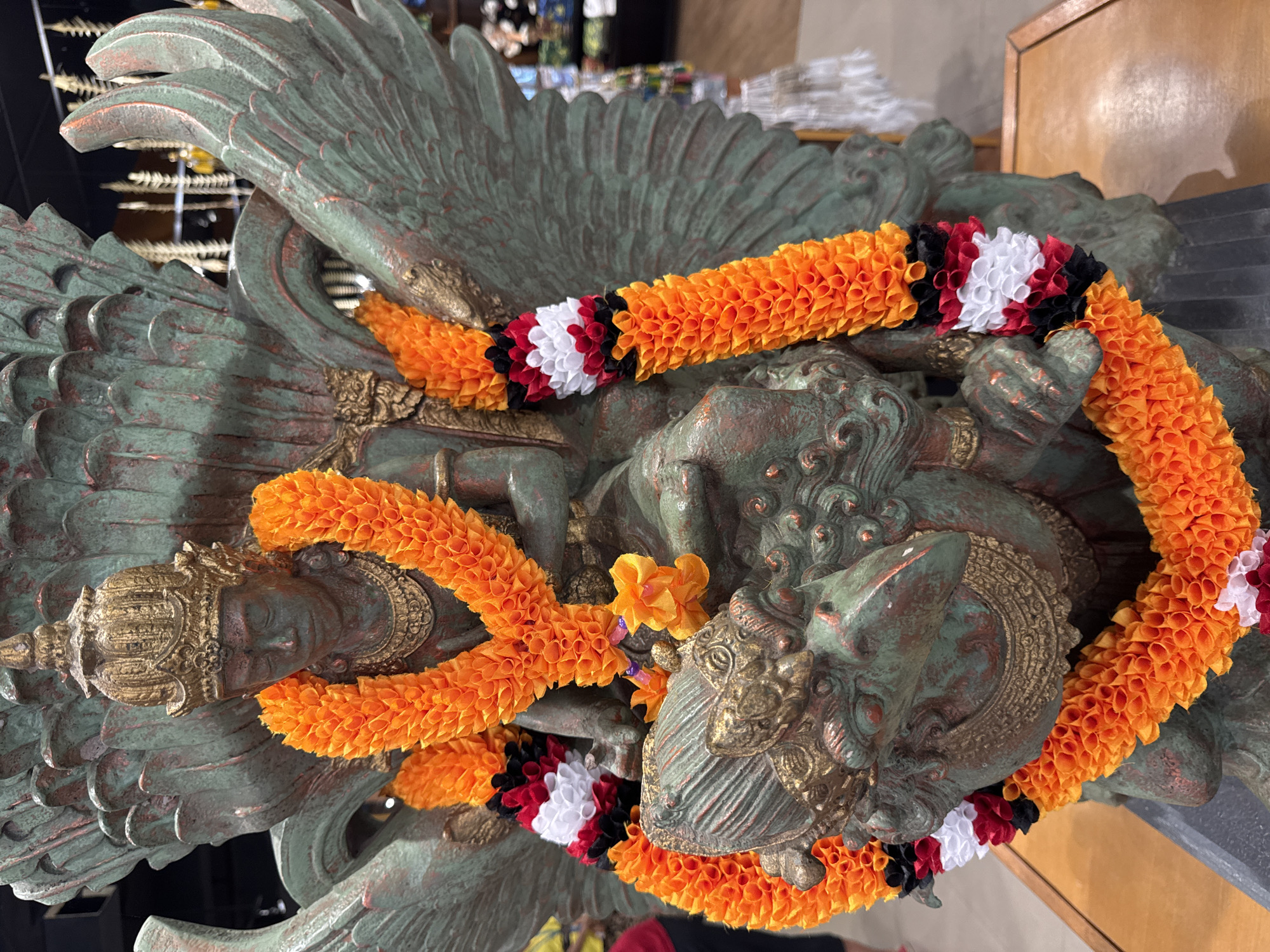 A statue with wings is adorned with an orange and black-white-red flower garland. The statue appears to be made of bronze and has a greenish patina. The location is Kuta Selatan, Indonesia.