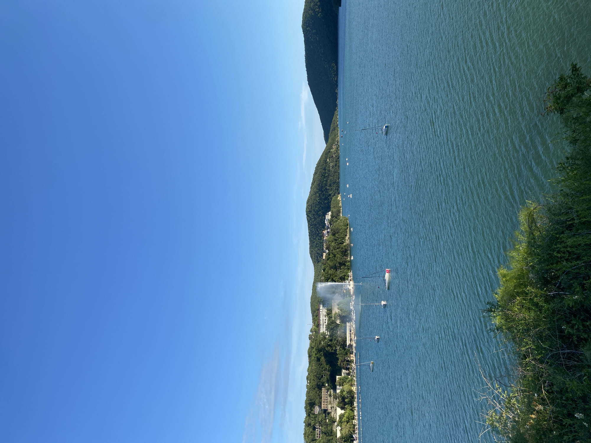 A wide-angle, eye-level shot captures a serene bay in Abrau-Dyurso, Russia, under a clear blue sky. The deep blue water of the bay is calm, with gentle ripples reflecting the bright sunlight. Several sailboats are scattered across the water, their white sails standing out against the azure backdrop. On the shore, lush green hills rise steeply, dotted with buildings that appear to be hotels or resorts. A prominent water fountain sprays a tall plume of water into the air near the coastline, creating a focal point. The foreground is occupied by dense green foliage, adding depth and texture to the scene. The overall atmosphere is one of tranquility and natural beauty, with a hint of human activity around the bay.