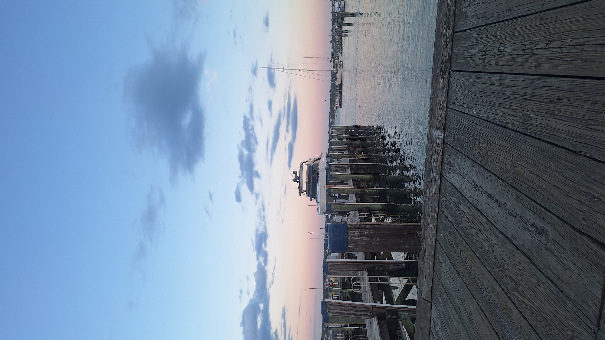 The image captures a serene harbor scene in Nantucket, United States of America, likely during twilight given the soft hues in the sky. In the foreground, a weathered wooden dock stretches out, its planks showing signs of age and use. Beyond the dock, a sleek, modern yacht is moored to a pier, its white hull gleaming against the dusky water. The pier itself is constructed of sturdy wooden pilings and planks, with metal ladders visible for access to the water.
Further out in the harbor, a tall-masted sailboat stands silhouetted against the horizon, its sails furled. The water is calm, reflecting the muted colors of the sky and the surrounding boats. Several other smaller boats and piers are visible in the distance, hinting at the active marine life of the harbor. The sky above is a gradient of soft blues and pinks, with a few scattered clouds adding texture and depth to the scene. There are no people visible in the image. The overall atmosphere is one of tranquility and peacefulness.