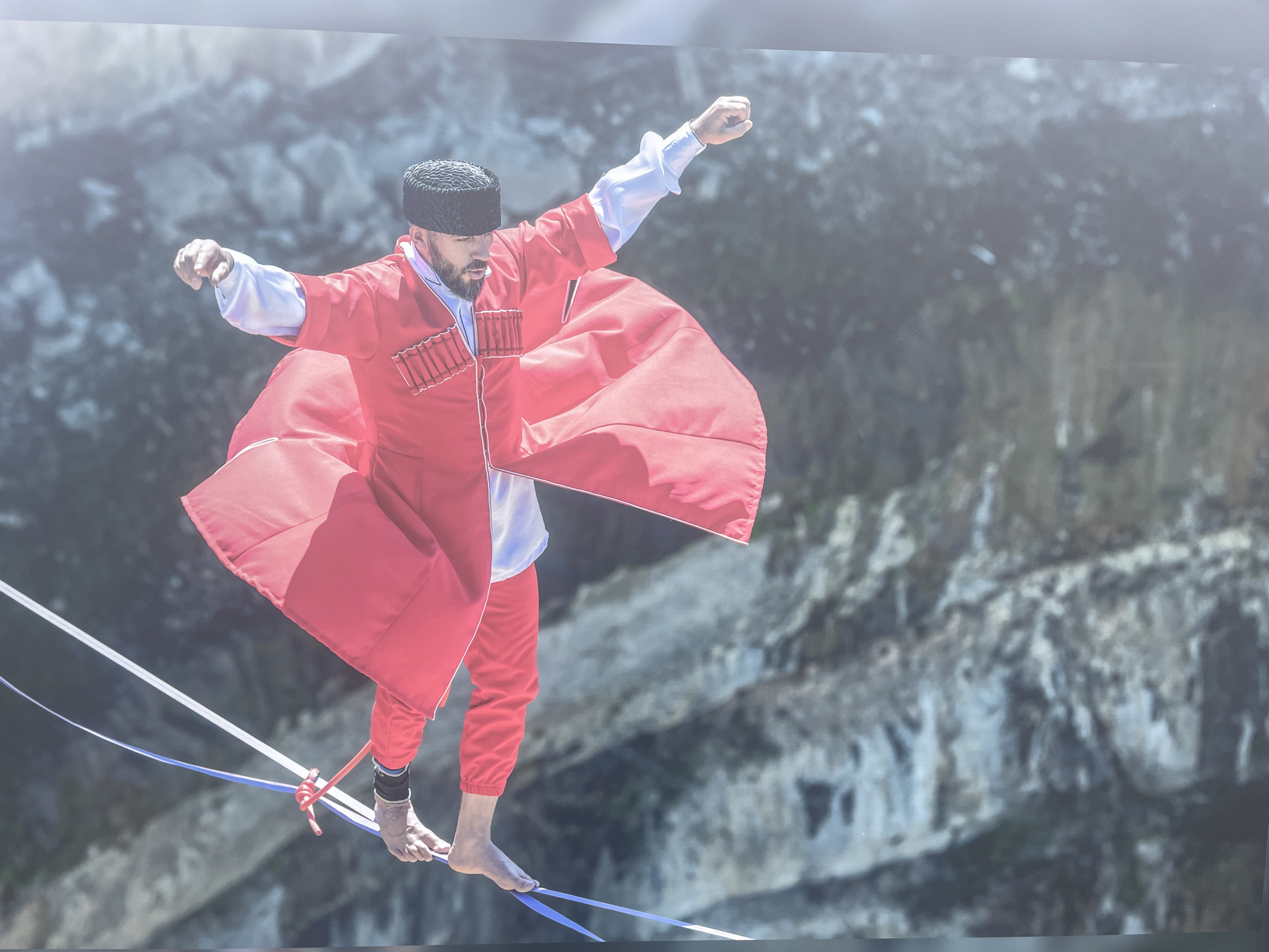 A man in traditional Caucasian attire performs a tightrope walk against a backdrop of rocky cliffs. He is wearing a red chokha with white detailing, a white shirt, and a black papakha. His arms are outstretched for balance as he walks barefoot across the rope. The setting appears to be in a mountainous region, possibly near Sochi, Russia. The backdrop is blurred due to the distance and possibly depth of field of the photograph.