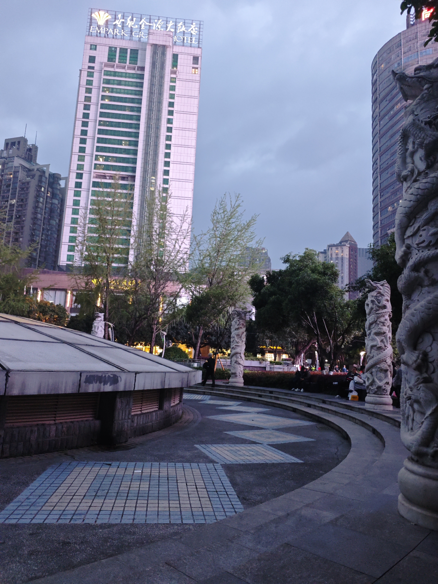 The image depicts a public space at dusk in Liangjiang, China. Dominating the background is the EmPark Grand Hotel, a tall, modern building with a lit sign at the top. Several other high-rise buildings are visible, including one to the right, featuring curved architecture. The foreground showcases a paved area with blue and white tiled patterns, bordered by low, curved steps. Several carved stone pillars with dragon designs are placed along the steps. People are gathered on the steps and in the background. The lighting suggests it is late evening.