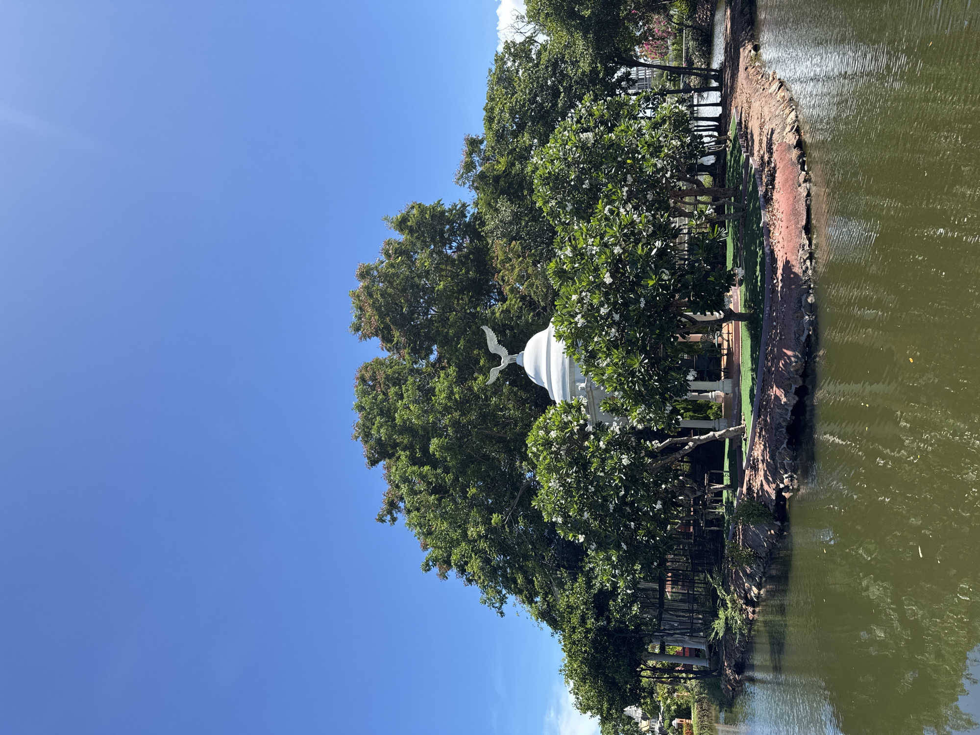 The image shows a partially obscured white pavilion with an eagle statue on top, set amidst trees and located on the edge of a body of water. The location is the Bang Pu Subdistrict Municipality, Thailand, evidenced by the typical Thai architecture style and local vegetation. A clear blue sky is overhead. The water shows some rippling and reflections, suggesting slight wind. Vegetation lines the water's edge, with trees of varying sizes including some with white blossoms. The pavilion's base is not fully visible due to the trees, but its general structure is evident. A small walkway or path runs along the water's edge in the vicinity of the pavilion. There are no people visible in the image.