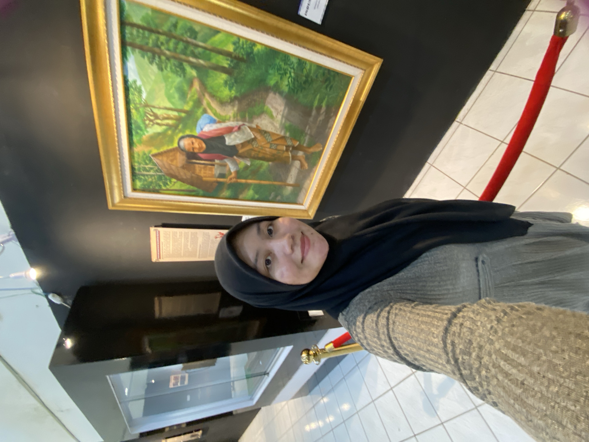 A woman wearing a black hijab and a grey sweater takes a selfie in a museum. She is standing in front of a large framed painting of an elderly woman walking on a dirt path in a lush, green landscape. A small, triangular hut is visible in the background of the painting. A red velvet rope barrier is visible in the foreground of the selfie. The museum has white tiled floors and black walls. The image appears to have been taken in the Museum Lambung Mangkurat in Banjarbaru, Indonesia.