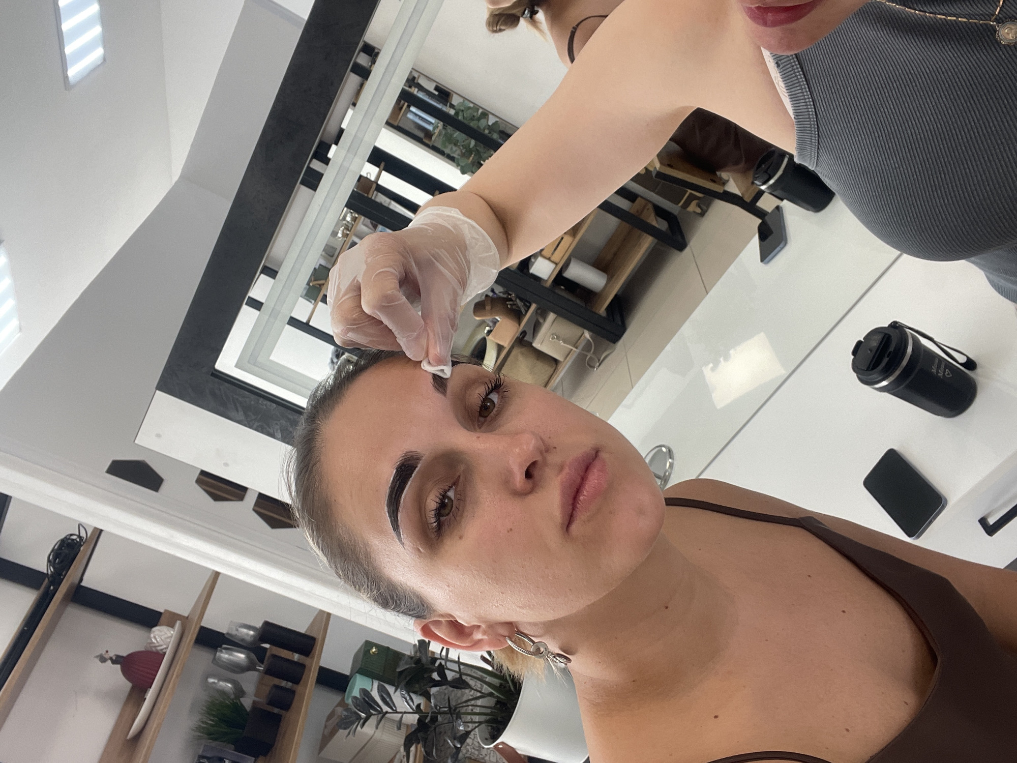 The photograph depicts a woman undergoing an eyebrow treatment in a beauty salon. The woman is lying down, facing upwards, with her head slightly tilted to the left. Her eyebrows are dark and well-defined, likely having just been treated. A gloved hand, presumably belonging to a beautician, is holding a tool near her right eyebrow. The beautician is partially visible, wearing a gray top.
In the background, there is a white table with a black water bottle and a black smartphone on it. The salon appears to be modern and well-lit, with shelves displaying various beauty products. A mirror reflects the interior of the salon, including a plant in a white pot.
Based on the information provided, the location is Chaikovsky, Russia.